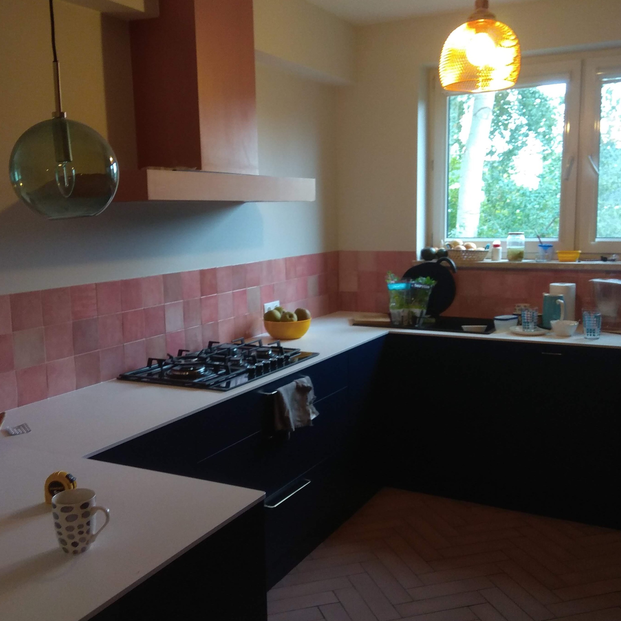 Nowoczesna kuchnia z ciemnymi szafkami, białym blatem i różowymi płytkami na ścianie. Na blacie widoczna płyta gazowa, miska z owocami, kubek w kropki i miarka. Podłoga wyłożona parkietem w jodełkę.