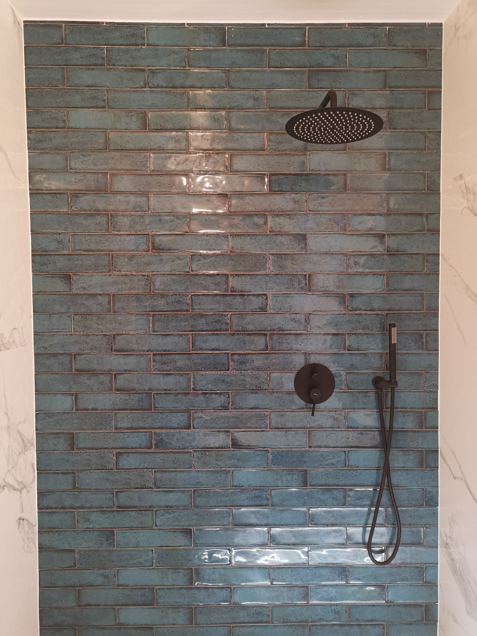 Ściana prysznicowa wyłożona błyszczącymi, turkusowymi płytkami w układzie cegiełki z czarną armaturą: deszczownica, bateria i słuchawka prysznicowa.