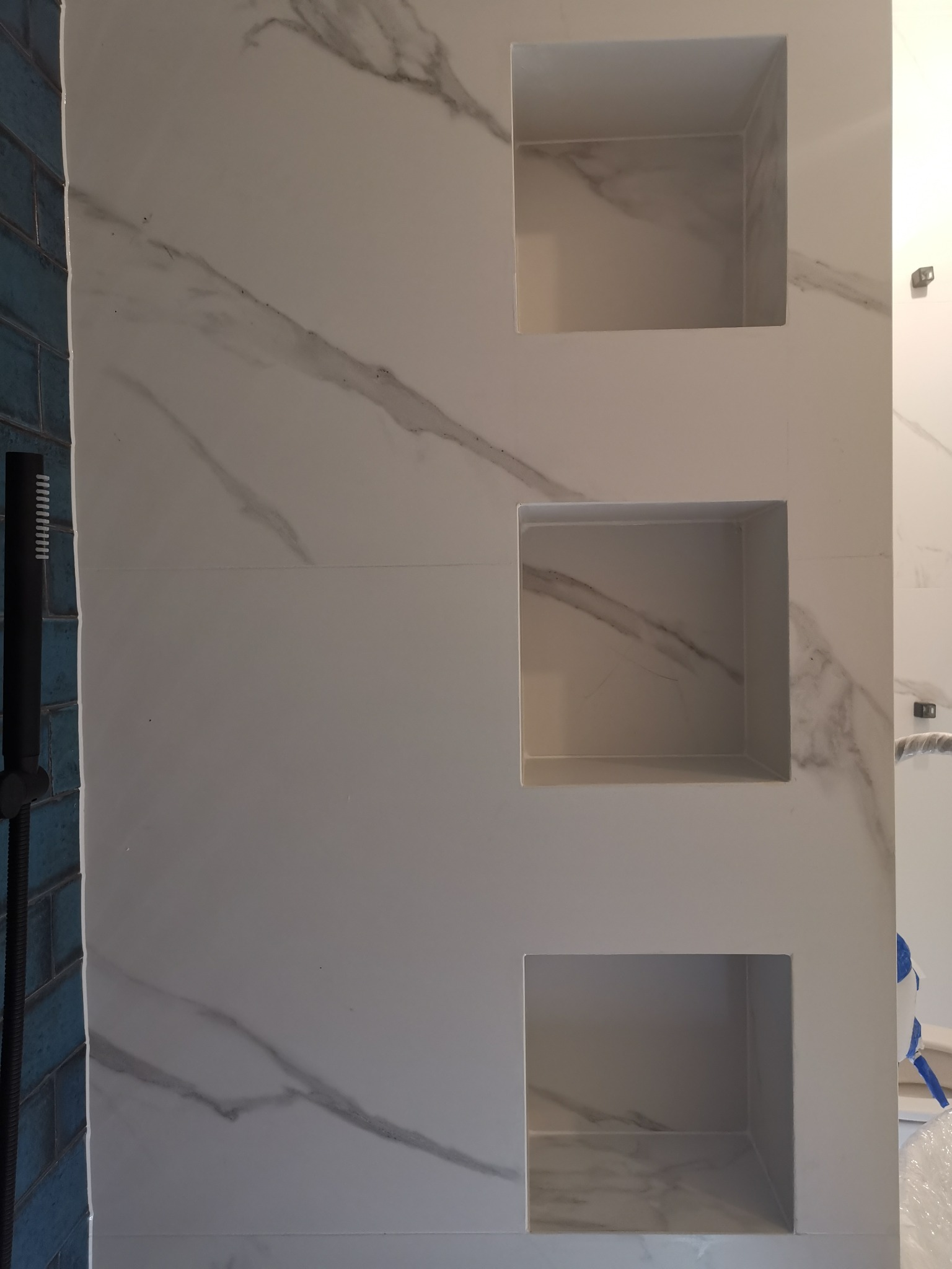 Ściana łazienki wyłożona płytkami imitującymi marmur z trzema wnękami, obok fragment niebieskich płytek i czarna słuchawka prysznicowa.