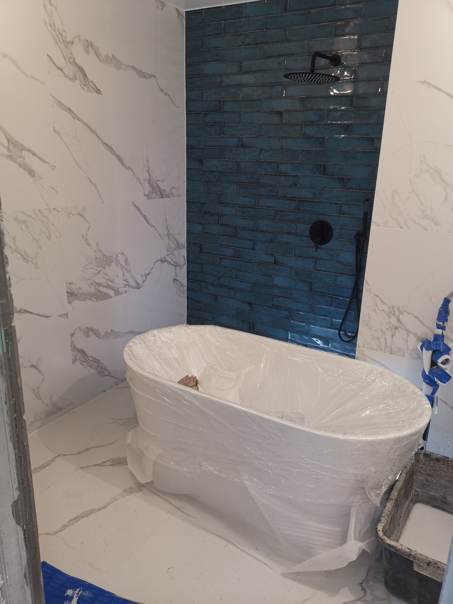 Nowa łazienka w trakcie wykańczania: wolnostojąca wanna zabezpieczona folią, ściany wyłożone białymi płytkami imitującymi marmur oraz niebieskimi, błyszczącymi płytkami cegiełkami, czarna...