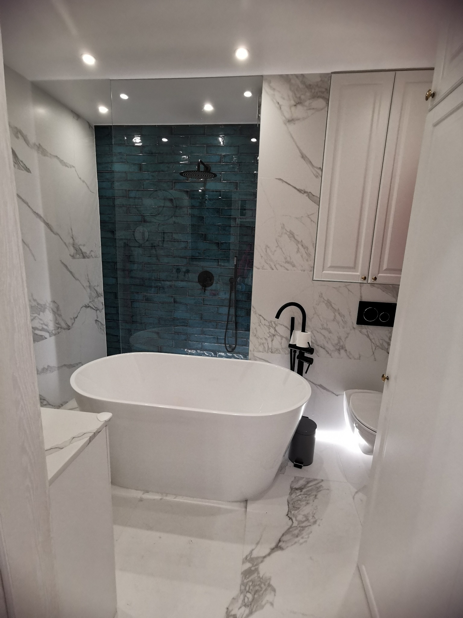 Nowoczesna łazienka z wolnostojącą wanną, kabiną prysznicową z turkusowymi płytkami i marmurowymi ścianami oraz podłogą, oświetlona punktowymi lampami sufitowymi.