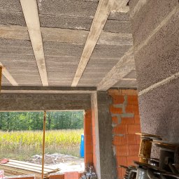 RD-West Bud - Wnętrze surowego domu w trakcie budowy: widok na strop z pustaków i belek, fragment ściany z cegły dziurawki, podpory stropowe i widok na zewnątrz przez otwór okienny.