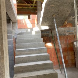 RD-West Bud - Surowe betonowe schody wewnątrz budynku w trakcie budowy, z widocznymi elementami konstrukcyjnymi i ceglanymi ścianami. W tle drewniany strop.