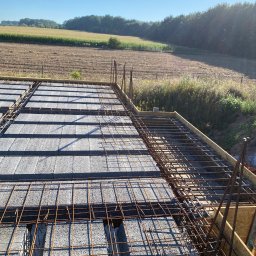 RD-West Bud - Zbrojenie stropu Teriva z widocznymi belkami, pustakami i stalową siatką zbrojeniową na tle krajobrazu polnych drzew w słoneczny dzień.