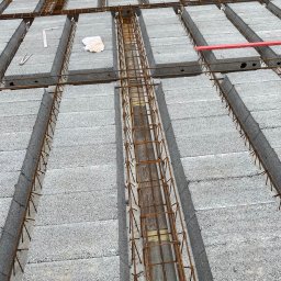 RD-West Bud - Szare bloczki stropowe zbrojone stalą, tworzące szkielet konstrukcji. Widoczne elementy drewniane i narzędzia budowlane. Ujęcie z góry, ukazujące detale zbrojenia.