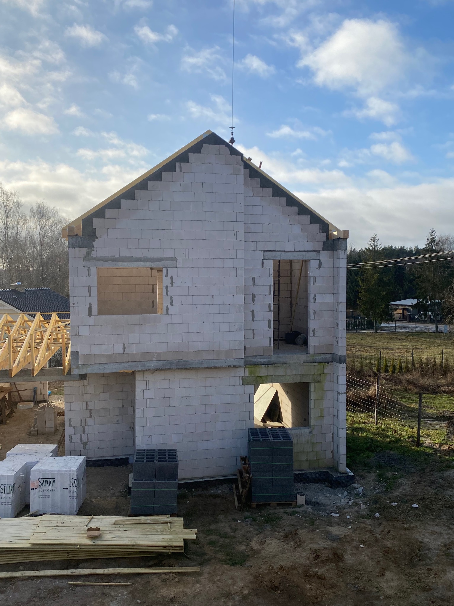 Surowy stan domu z białych bloczków, widoczne otwory okienne i drzwiowe, drewniana konstrukcja dachu w tle, materiały budowlane na pierwszym planie, słoneczny dzień.