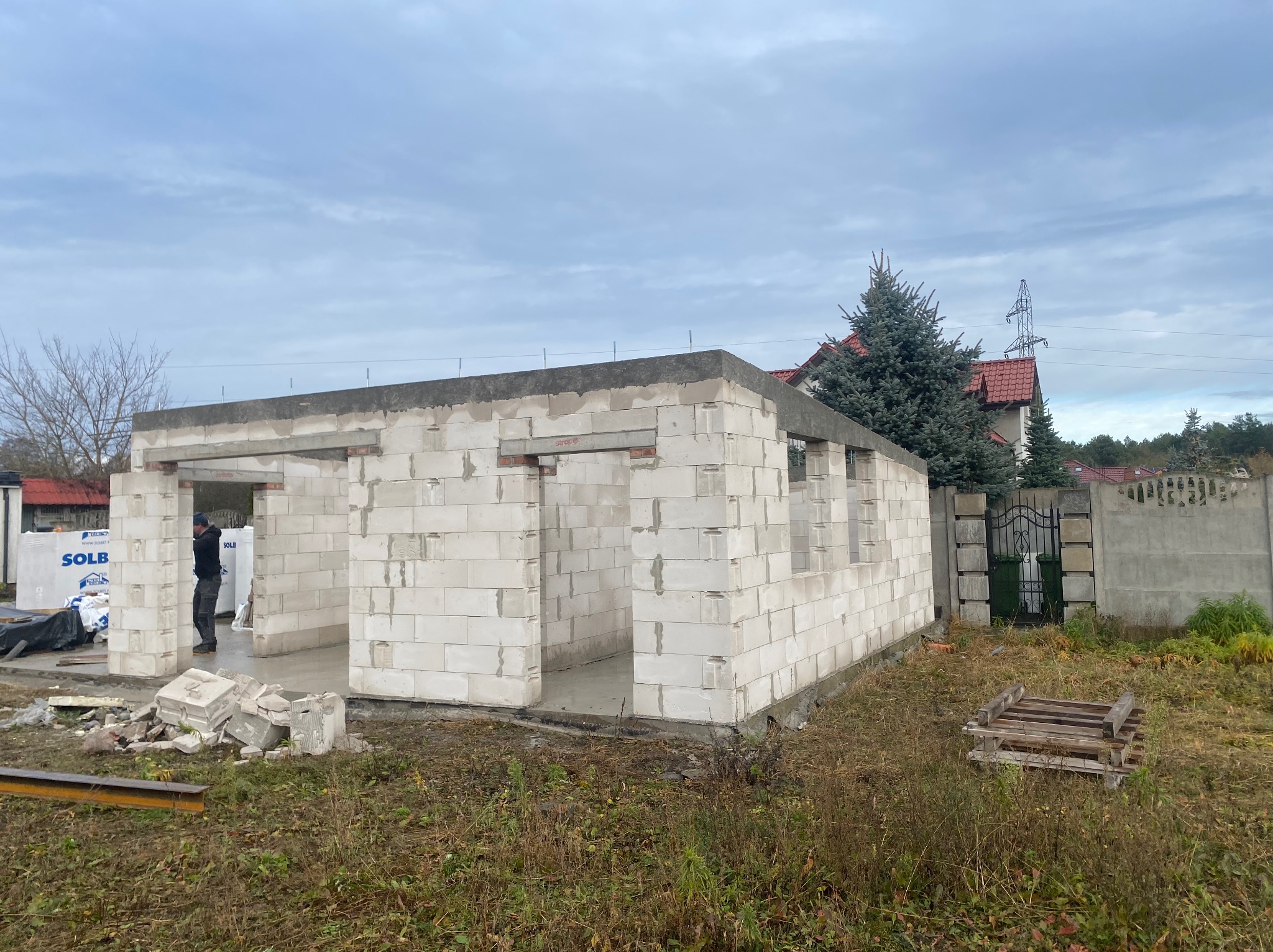 Budowa garażu z betonu komórkowego: ściany wzniesione, widoczne nadproża, teren budowy z gruzem i paletą, pracownik w tle. Stan surowy otwarty.