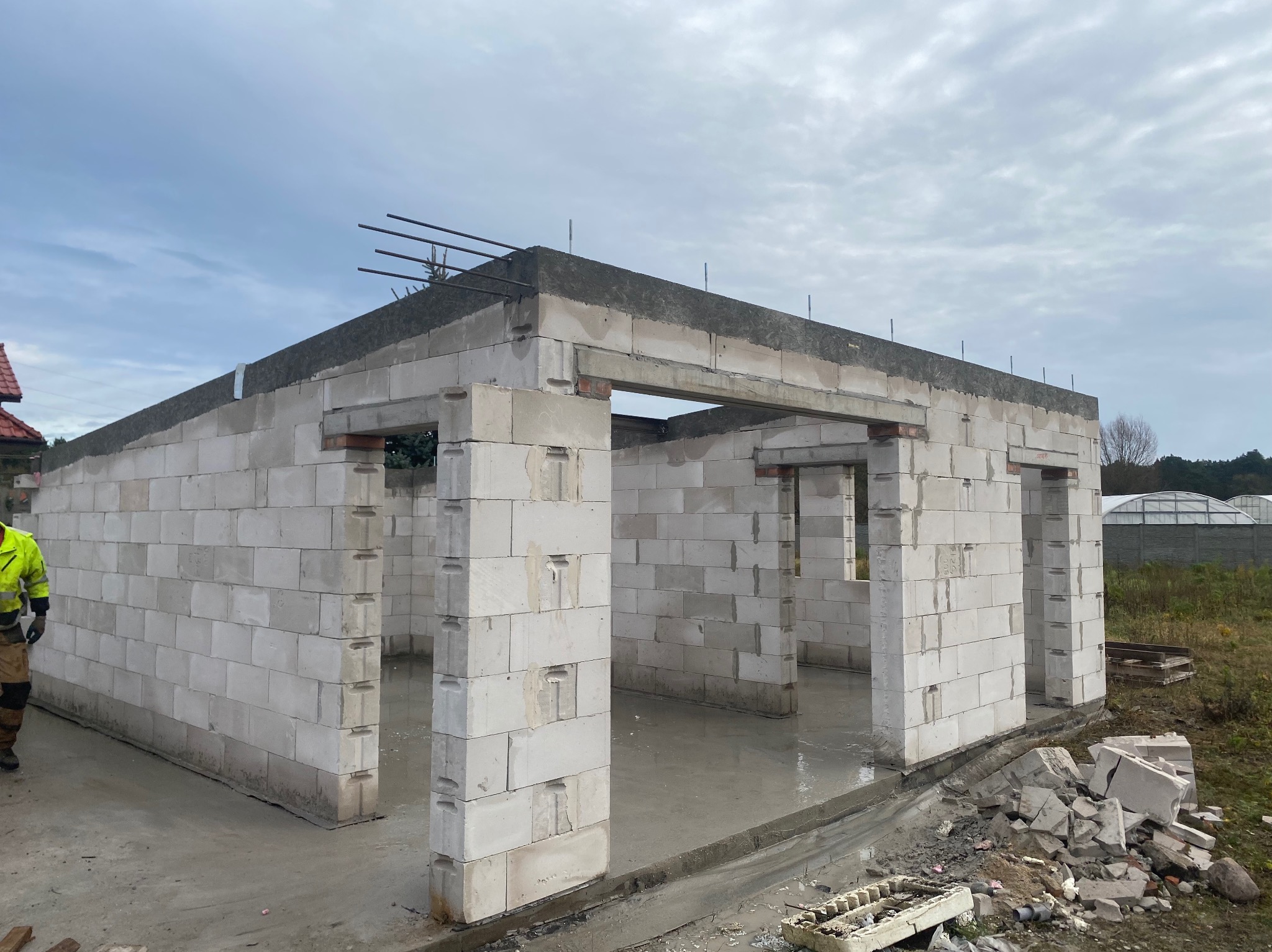 Wznoszenie ścian parterowego domu z betonu komórkowego, widoczne zbrojenie wieńca. Na pierwszym planie resztki materiałów budowlanych, w tle zabudowa jednorodzinna.