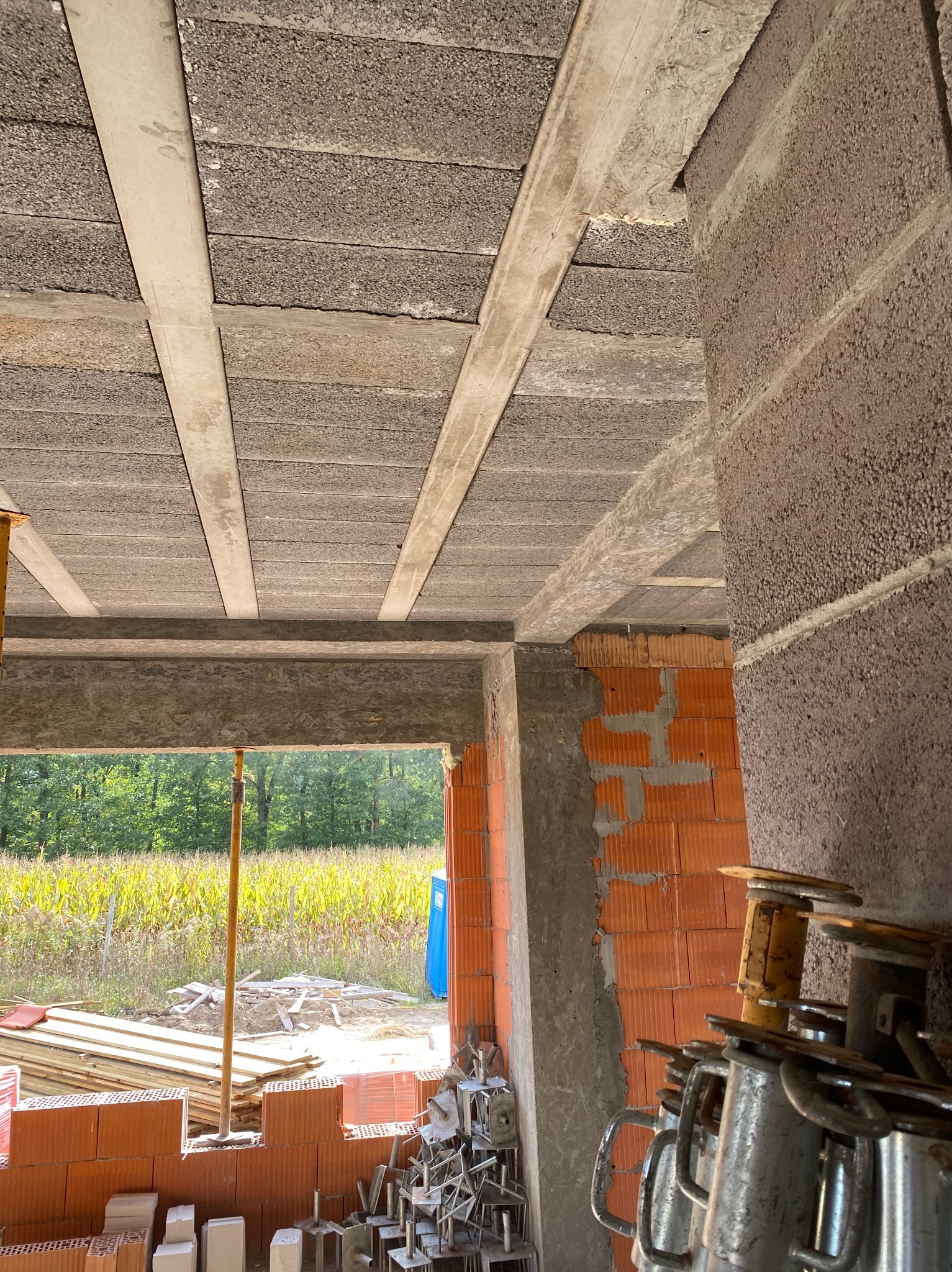 Wnętrze surowego domu w trakcie budowy: widok na strop z pustaków i belek, fragment ściany z cegły dziurawki, podpory stropowe i widok na zewnątrz przez otwór okienny.