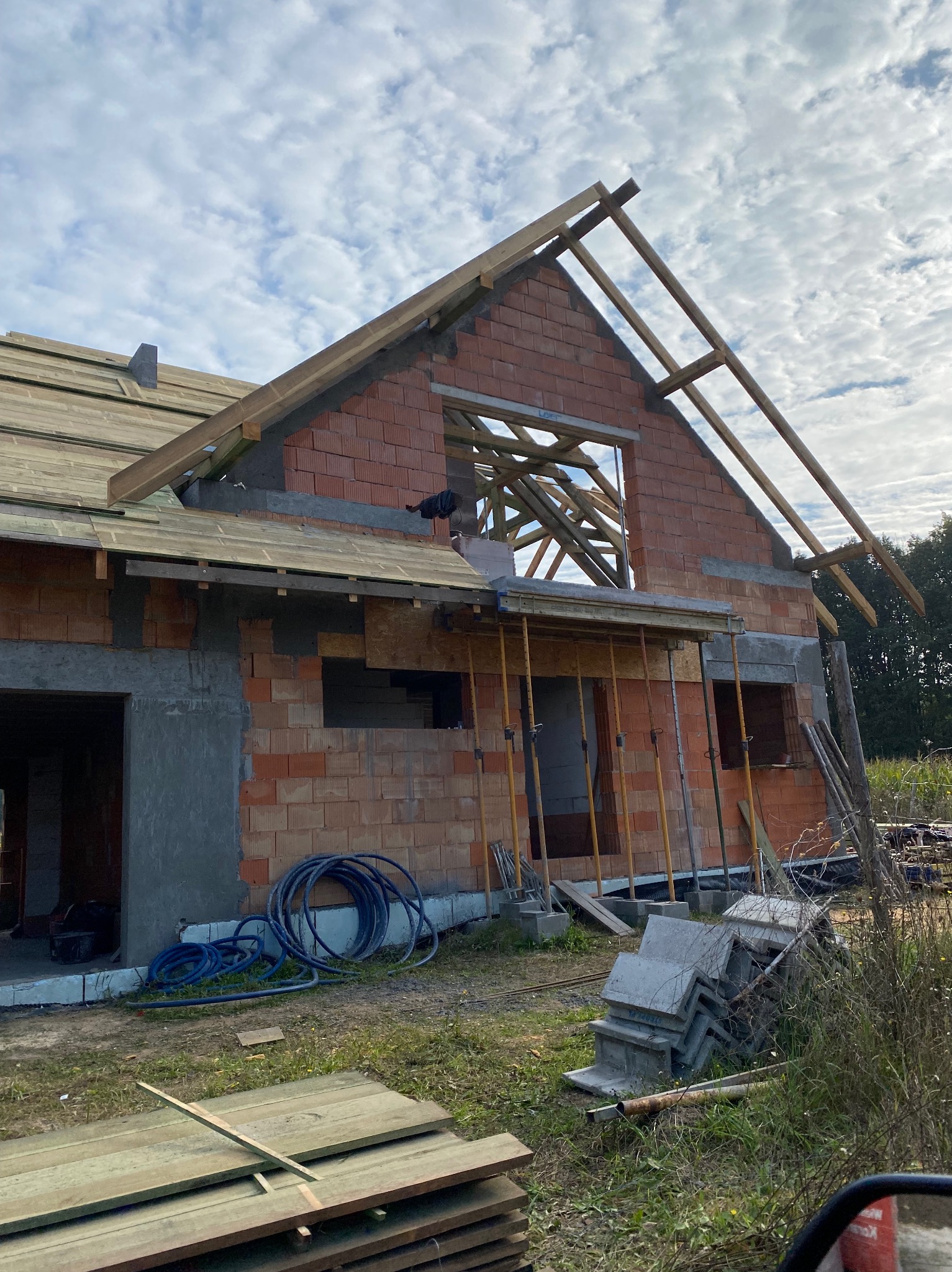 Budowa domu z czerwonej cegły w trakcie realizacji, widoczne drewniane elementy konstrukcji dachu i rusztowania. Na pierwszym planie deski i węże budowlane.