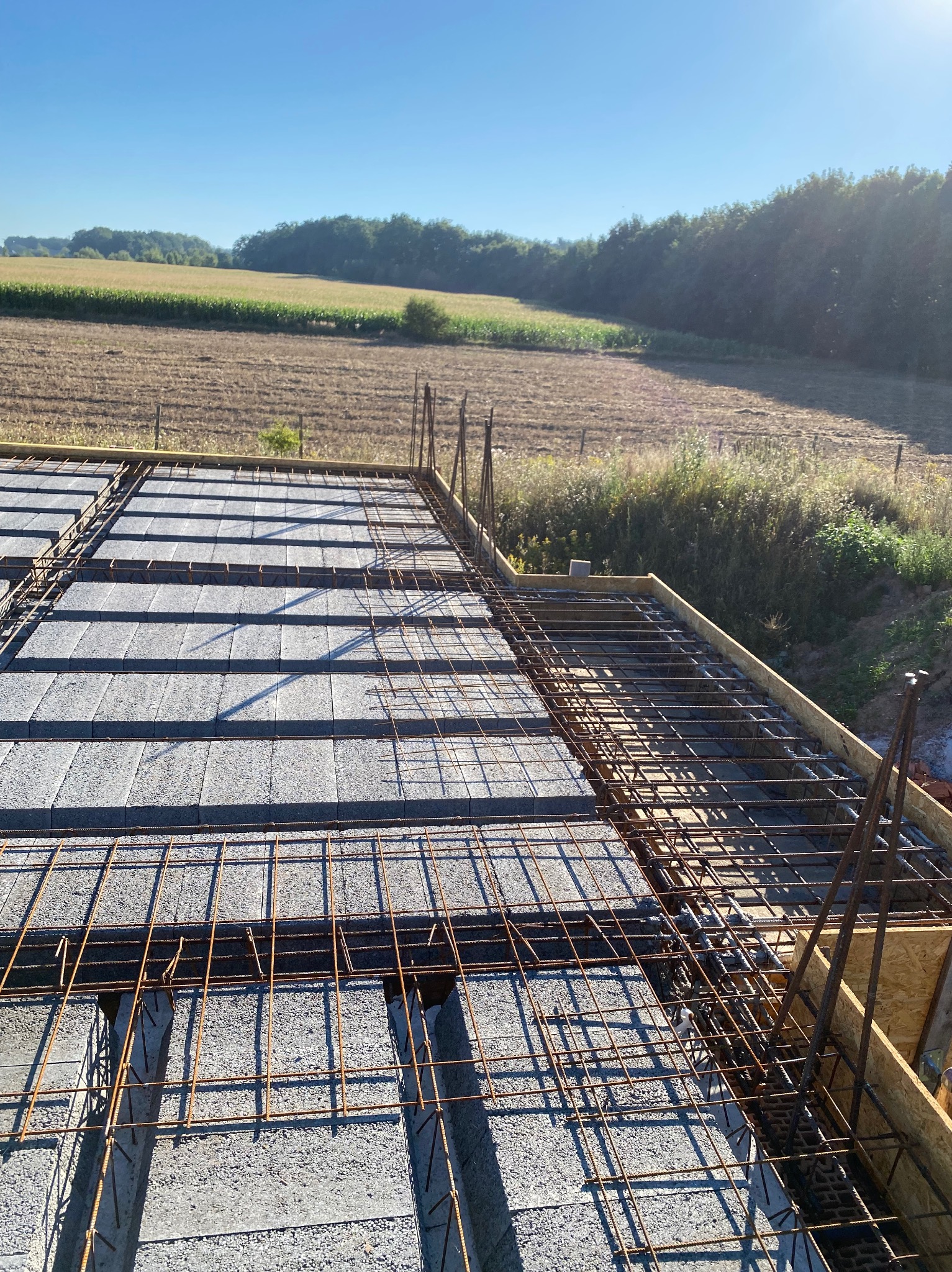 Zbrojenie stropu Teriva z widocznymi belkami, pustakami i stalową siatką zbrojeniową na tle krajobrazu polnych drzew w słoneczny dzień.
