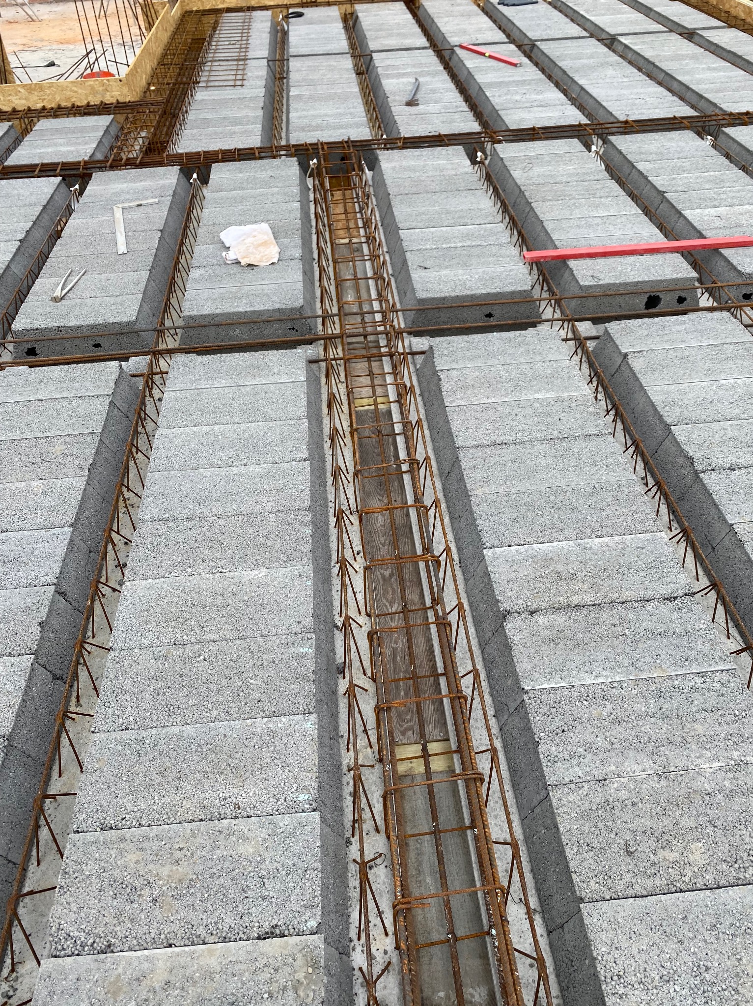 Szare bloczki stropowe zbrojone stalą, tworzące szkielet konstrukcji. Widoczne elementy drewniane i narzędzia budowlane. Ujęcie z góry, ukazujące detale zbrojenia.