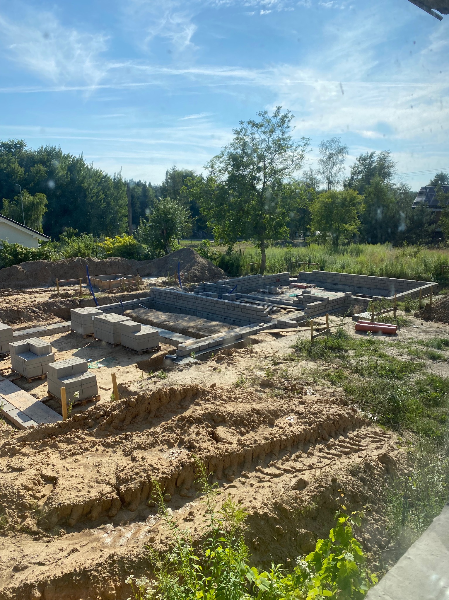 Wykop pod fundamenty domu z częściowo wymurowanymi ścianami z bloczków betonowych. Widoczne palety z bloczkami i przygotowany teren budowy w słoneczny dzień.