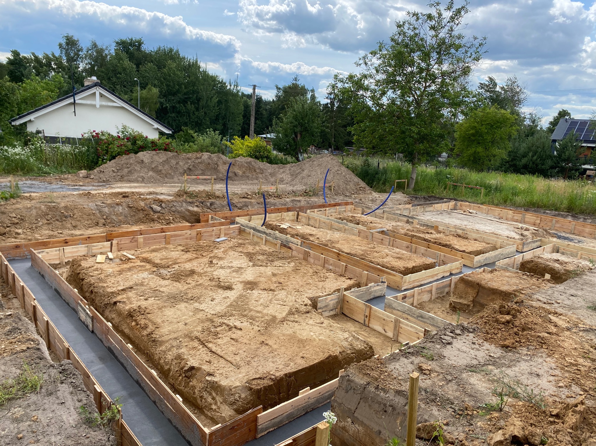 Wykopane fundamenty z drewnianym szalunkiem i częściowo wylanym betonem, widok z góry na plac budowy z domem w tle i błękitnymi rurami.