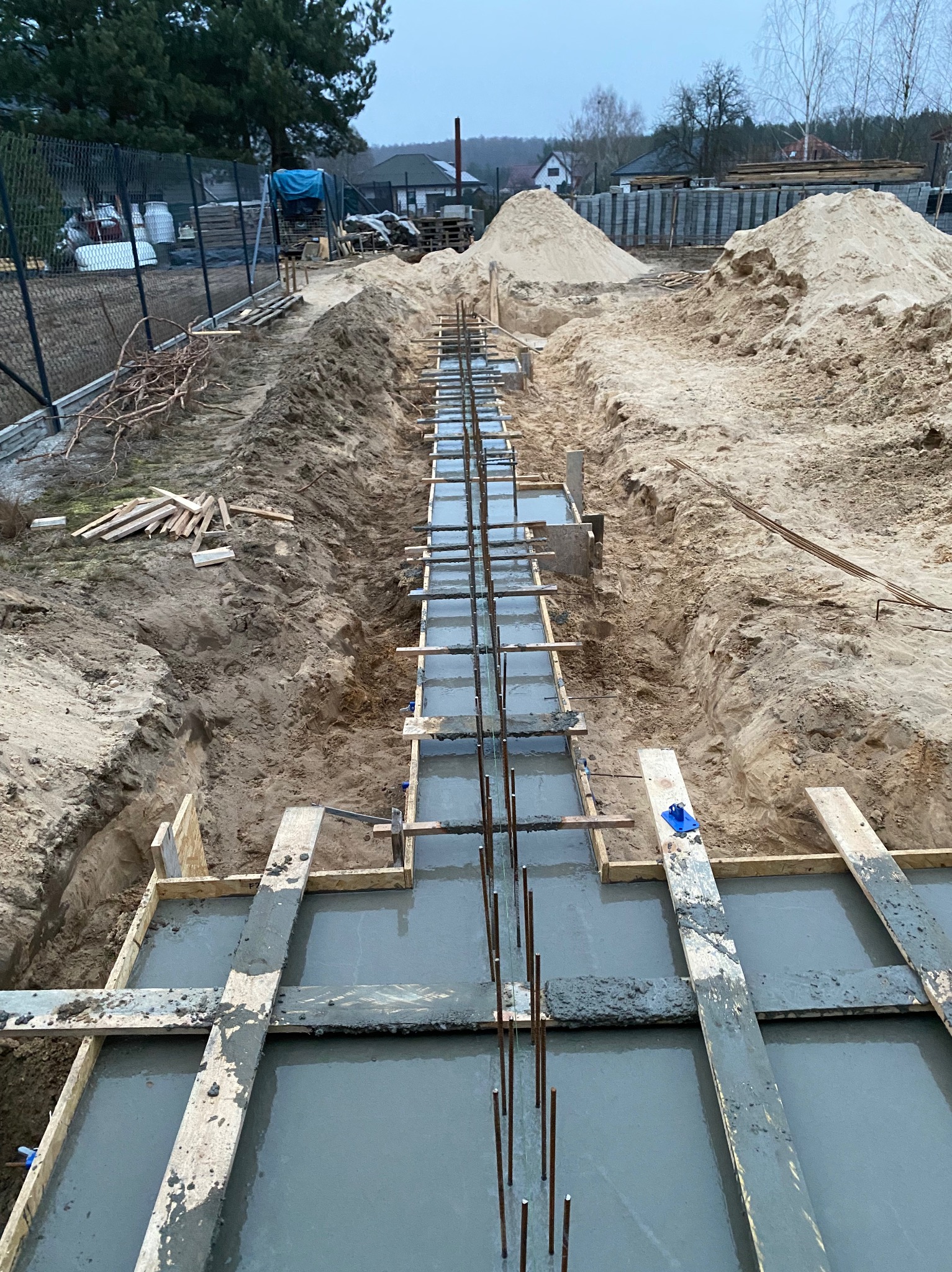 Wykop pod fundamenty zalany betonem, z widocznym zbrojeniem i drewnianym szalunkiem. W tle hałda piasku i zabudowa jednorodzinna. Ujęcie z góry.