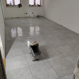 Metr po metrze - Ułożone szare płytki podłogowe w jasnym pomieszczeniu, widoczny wózek glazurniczy i okna z parapetami. Świeżo położona podłoga, pomieszczenie w trakcie prac wykończeniowych.