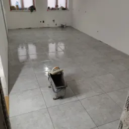 Ułożone szare płytki podłogowe w jasnym pomieszczeniu, widoczny wózek glazurniczy i okna z parapetami. Świeżo położona podłoga, pomieszczenie w trakcie prac wykończeniowych.