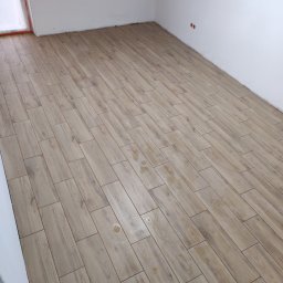 Metr po metrze - Ukończona podłoga z płytek imitujących drewno, ułożonych diagonalnie w jasnym pomieszczeniu z białymi ścianami. Widoczne ślady po fugowaniu.