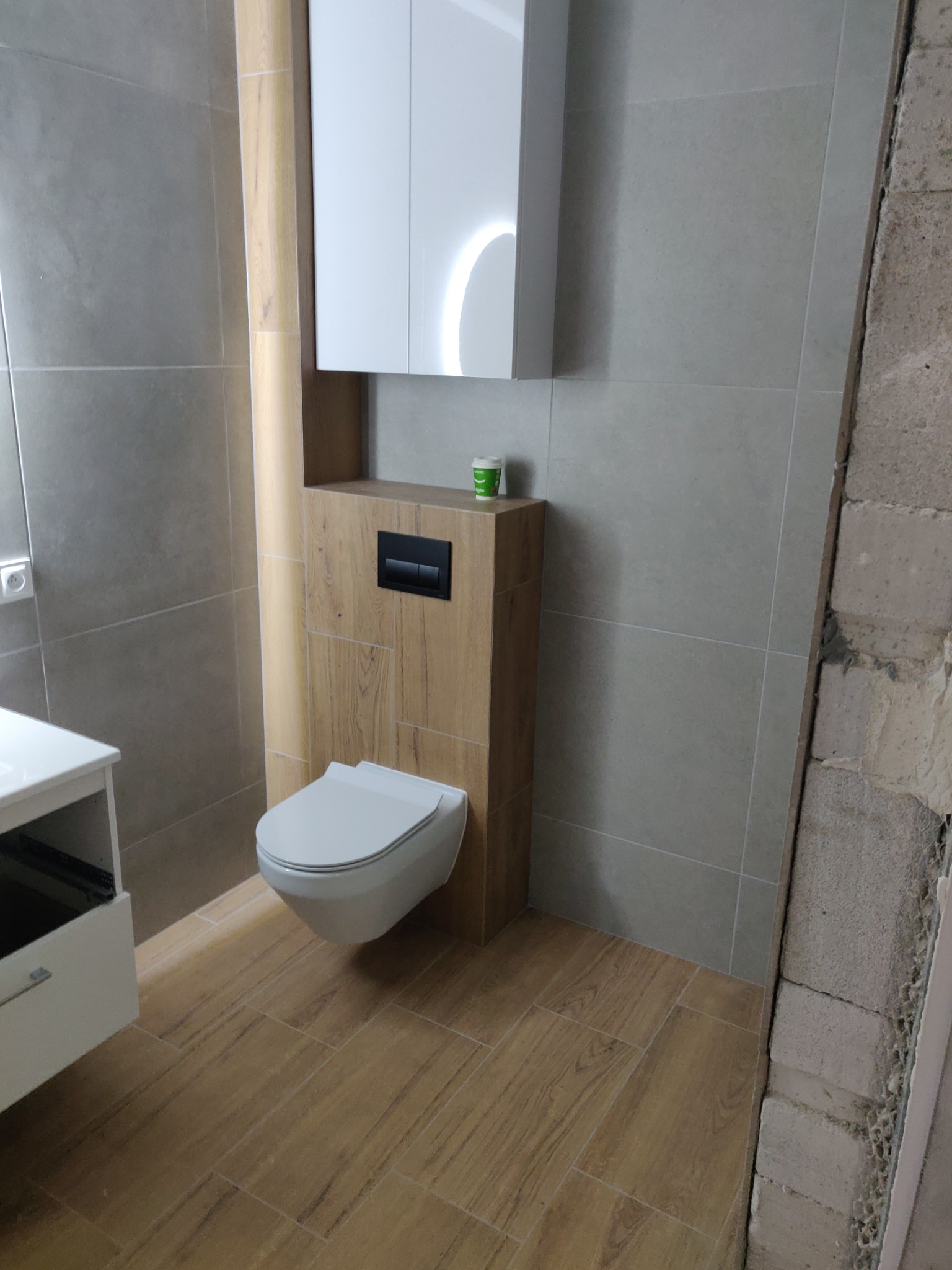 Nowoczesna łazienka po remoncie: podwieszana toaleta z zabudową w drewnie, szare płytki ścienne i podłogowe imitujące drewno. Elementy białej szafki i fragment ściany w surowym stanie.