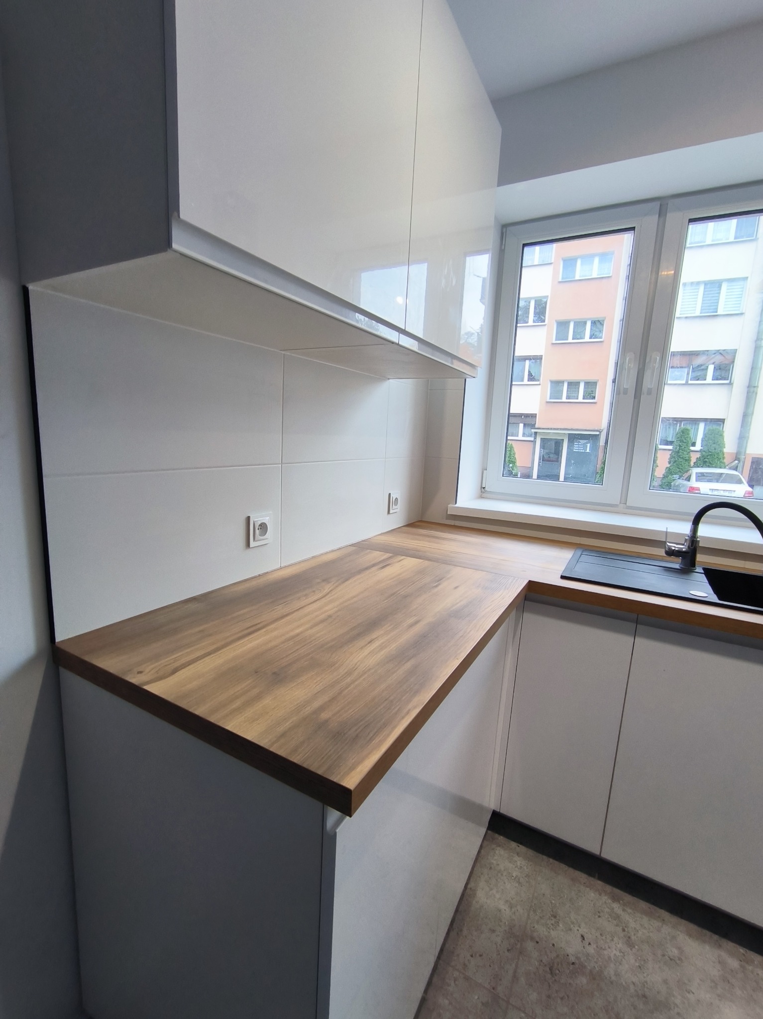 Nowoczesna kuchnia z białymi szafkami i drewnianym blatem. Widoczne gniazdka elektryczne na ścianie z białych płytek. Minimalistyczny design, okno z widokiem na budynek.