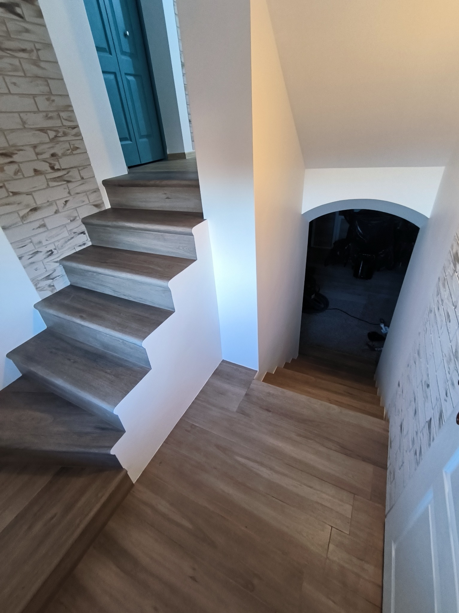 Wykończone schody z drewnopodobnymi panelami, białe ściany i niebieskie drzwi. Widok z góry na dół, z widocznym wejściem do piwnicy.