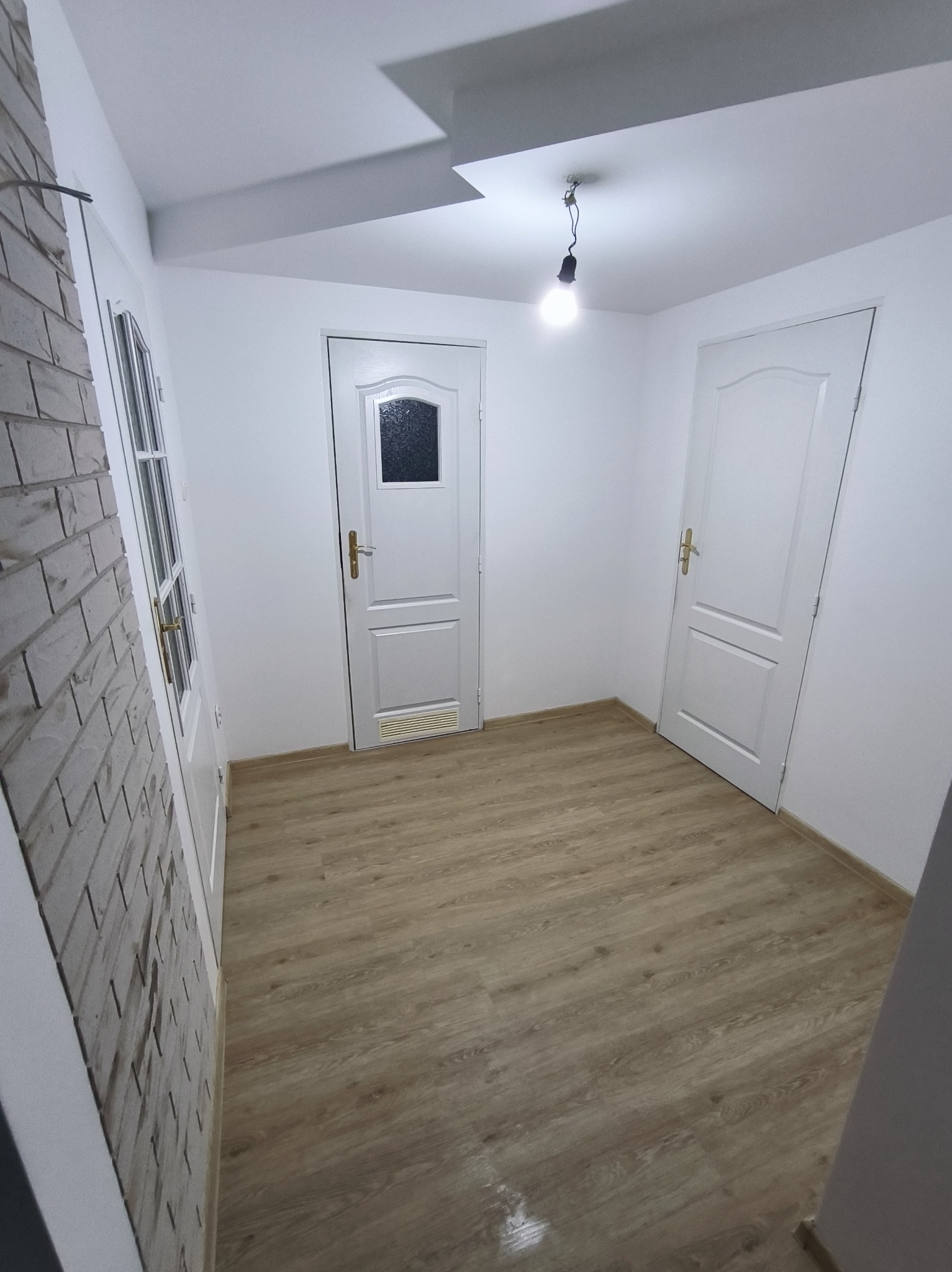 Wykończony korytarz w Koninie z panelami podłogowymi, białą cegłą na ścianie i dwiema białymi drzwiami. Widoczna żarówka. Minimalistyczny design.