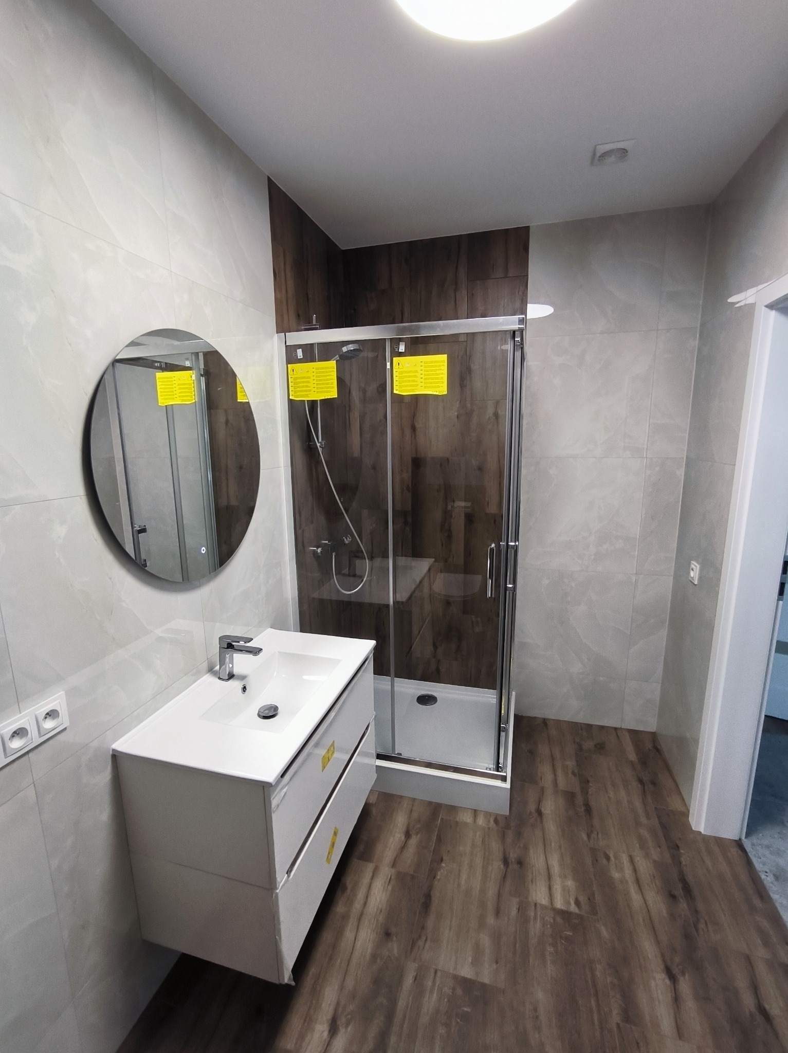 Nowoczesna łazienka z kabiną prysznicową, umywalką z szafką i okrągłym lustrem. Jasne płytki na ścianach, drewnopodobna podłoga. Minimalistyczny design.