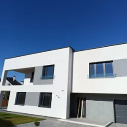 Nowoczesny dom z białą elewacją, szarymi panelami dekoracyjnymi, dużymi oknami z balustradami, zadbanym trawnikiem i wybrukowanym podjazdem pod błękitnym niebem.