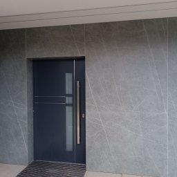 Vlodek Duzhak - Nowoczesne, ciemnoszare drzwi wejściowe z długą klamką i poziomymi listwami, osadzone w ścianie z jasnoszarych płytek o marmurowym żyłkowaniu. Przed drzwiami ciemna wycieraczka.