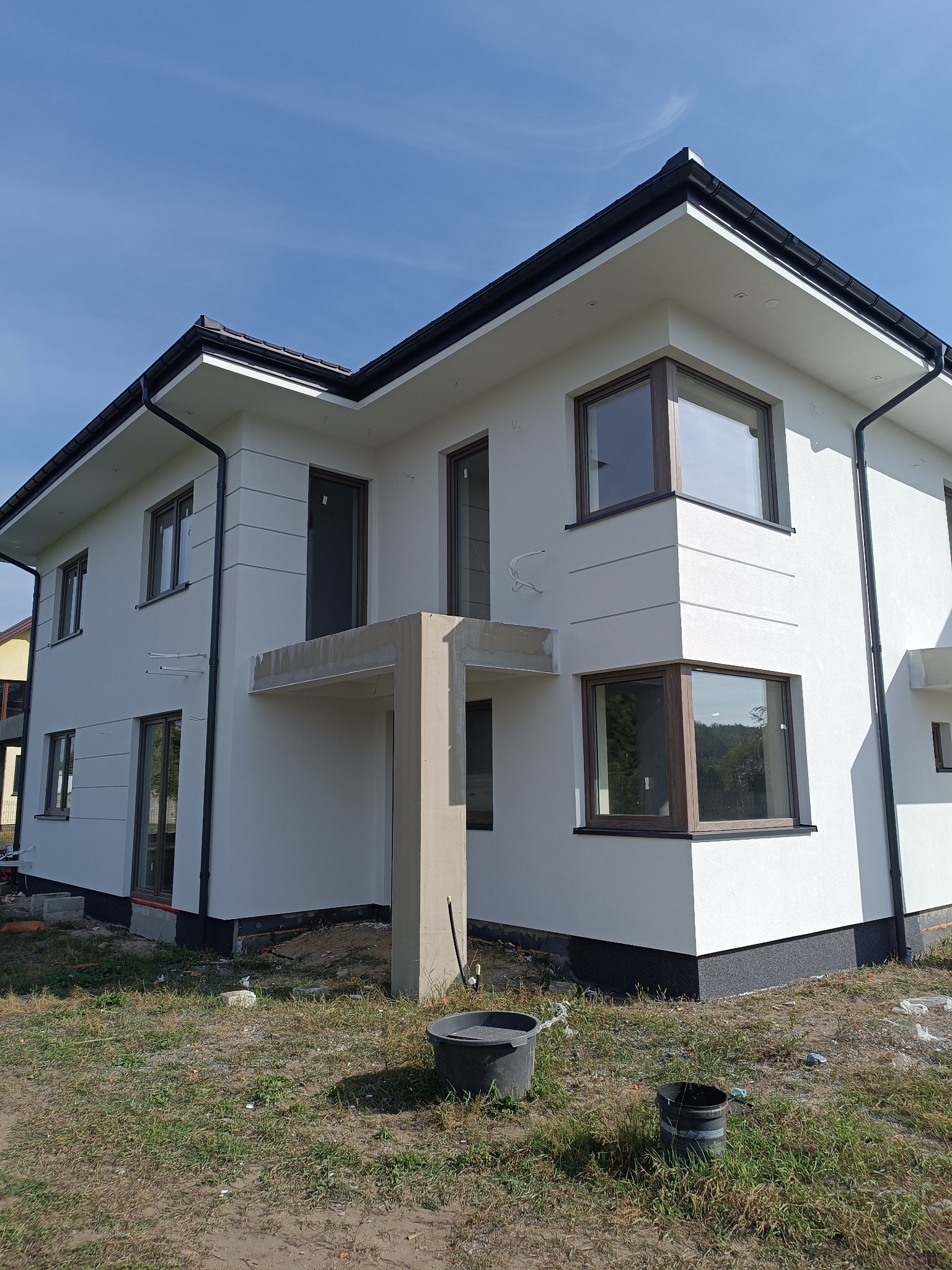 Nowoczesny, dwukondygnacyjny dom z białą elewacją i ciemnymi obramowaniami okien, widoczne detale wykończeniowe, czarne rynny, surowy stan otoczenia z widocznymi wiadrami i resztkami materiałów...