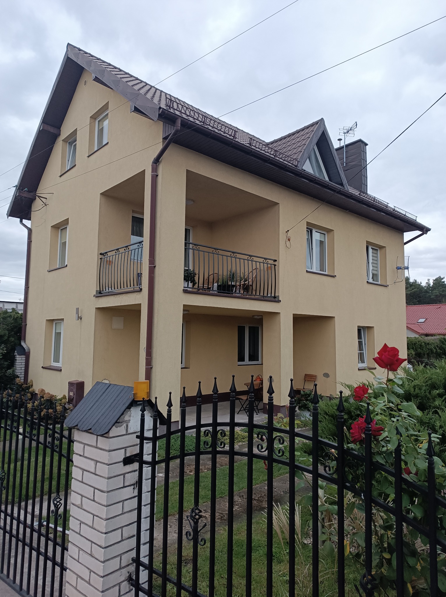 Dwupiętrowy dom z beżową elewacją, balkonami z czarnymi balustradami, widoczny zza czarnego, kutego ogrodzenia z kwitnącymi czerwonymi różami na pierwszym planie.