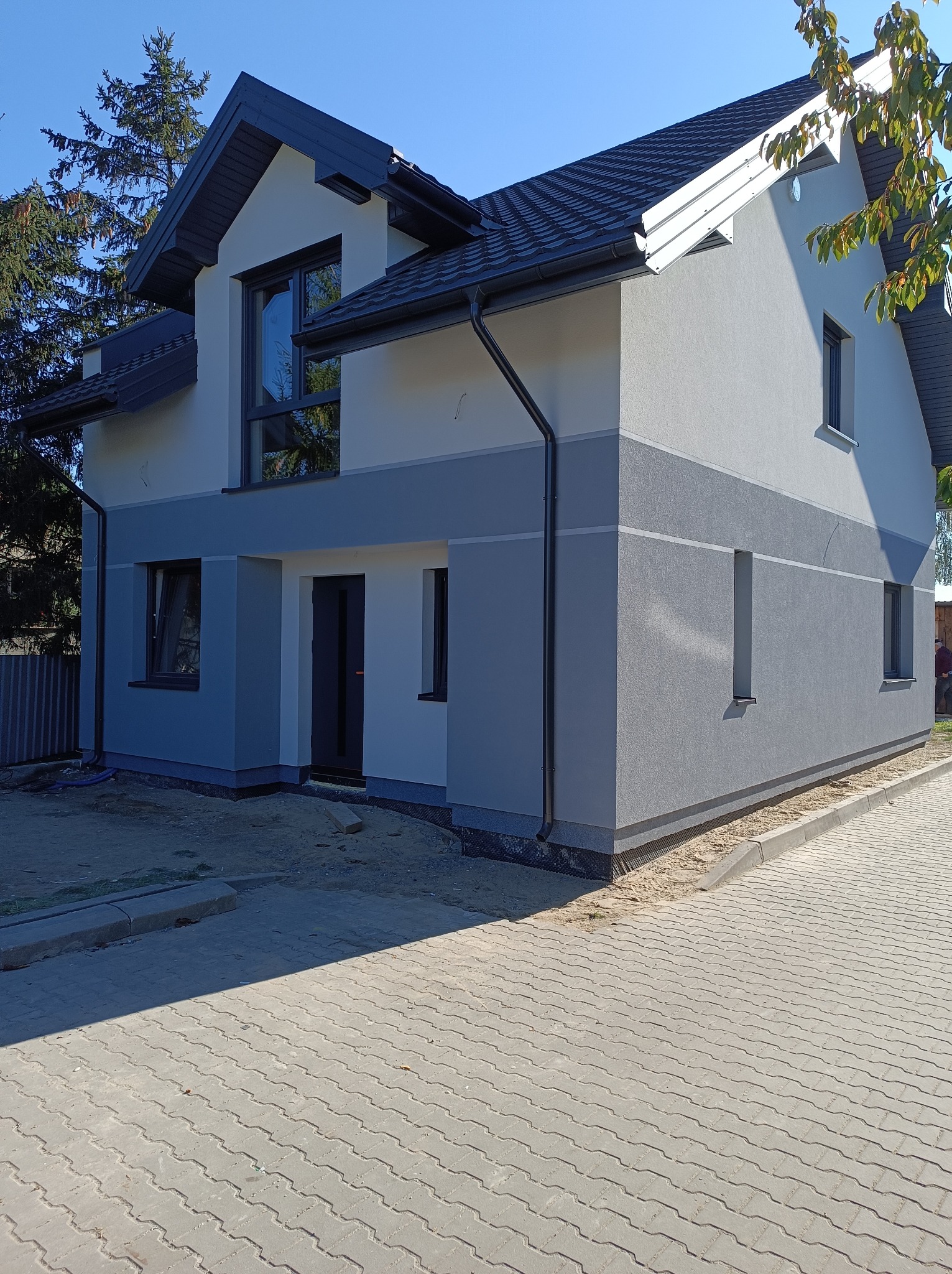 Nowoczesny dom jednorodzinny z dwukolorową elewacją w odcieniach szarości, ciemna stolarka okienna i drzwiowa, widoczne orynnowanie i kostka brukowa przed budynkiem.