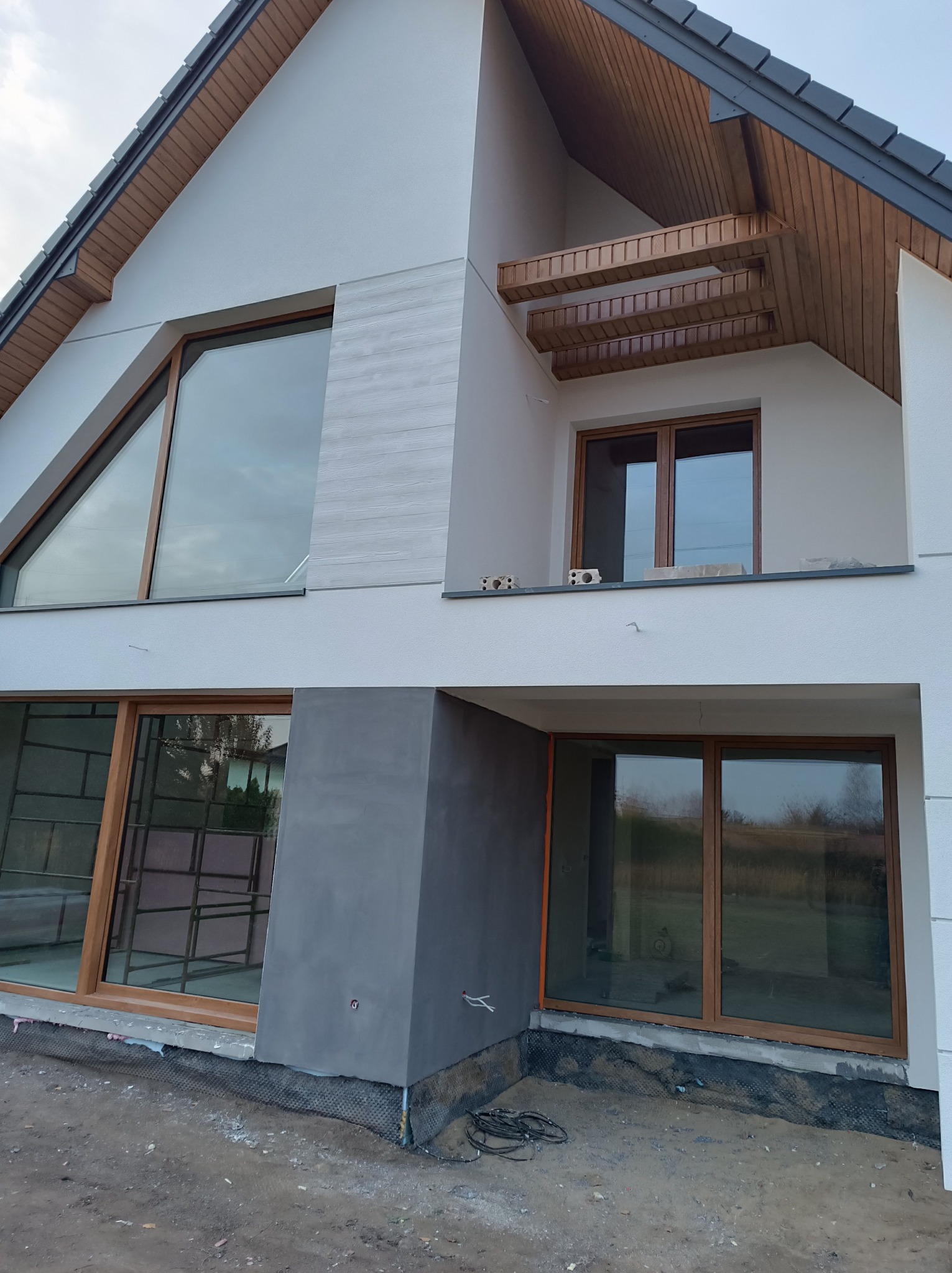 Nowoczesny dom jednorodzinny w trakcie prac elewacyjnych: widoczne fragmenty ocieplenia, tynkowane szare elementy oraz duże okna z drewnianą stolarką.