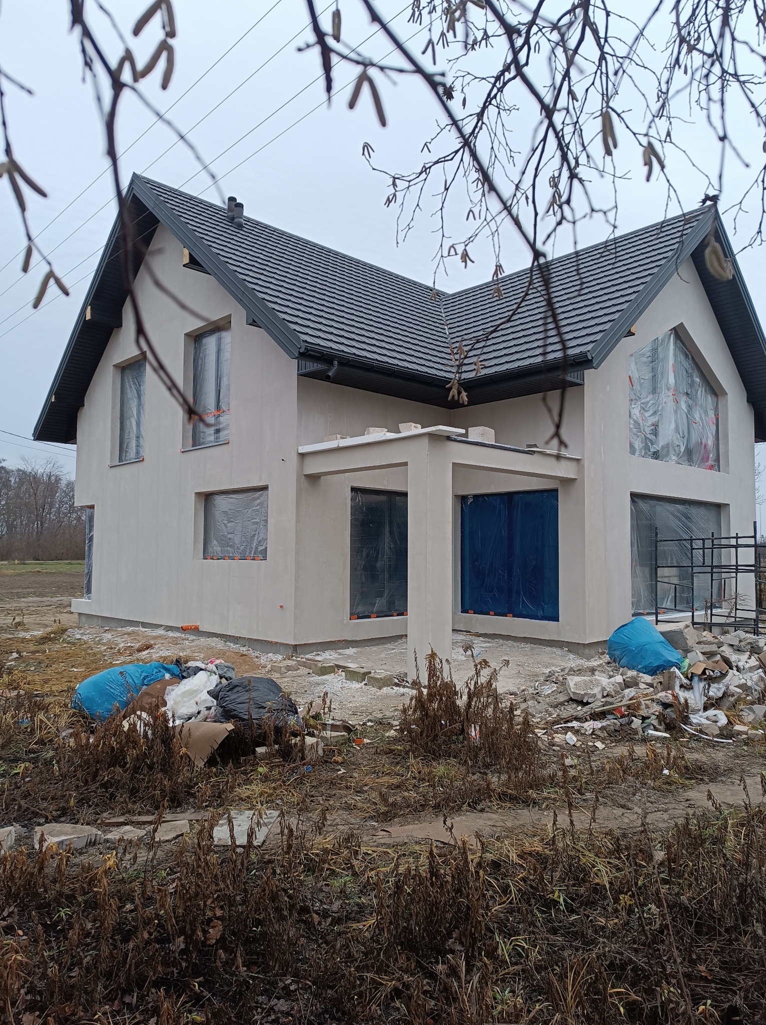 Dom jednorodzinny w trakcie prac wykończeniowych elewacji, okna zabezpieczone folią, widoczne ślady prac budowlanych i gruz na posesji.
