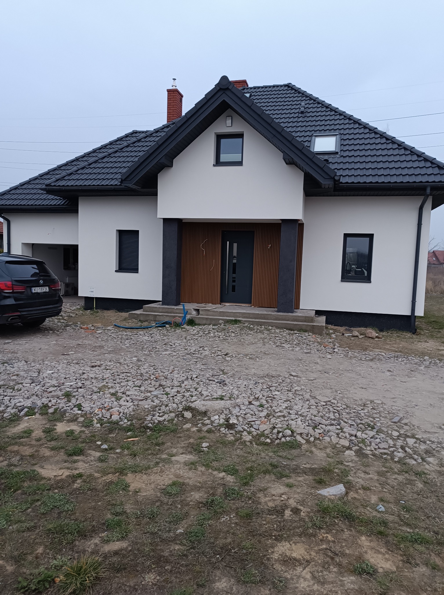 Nowoczesny dom jednorodzinny z białą elewacją, ciemną stolarką okienną i drewnianymi panelami przy wejściu, widok z zewnątrz na tle szarego nieba i gruzowiska.