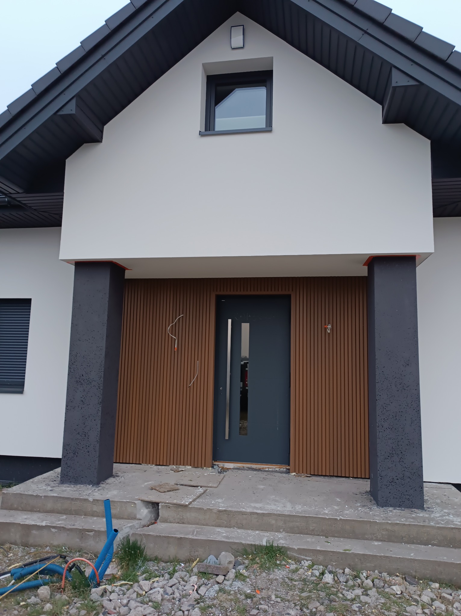 Nowoczesny dom jednorodzinny z elewacją w kolorze białym i drewnianymi panelami pionowymi przy wejściu, ciemnoszare kolumny i drzwi, widoczne niedokończone schody wejściowe i wystające kable.
