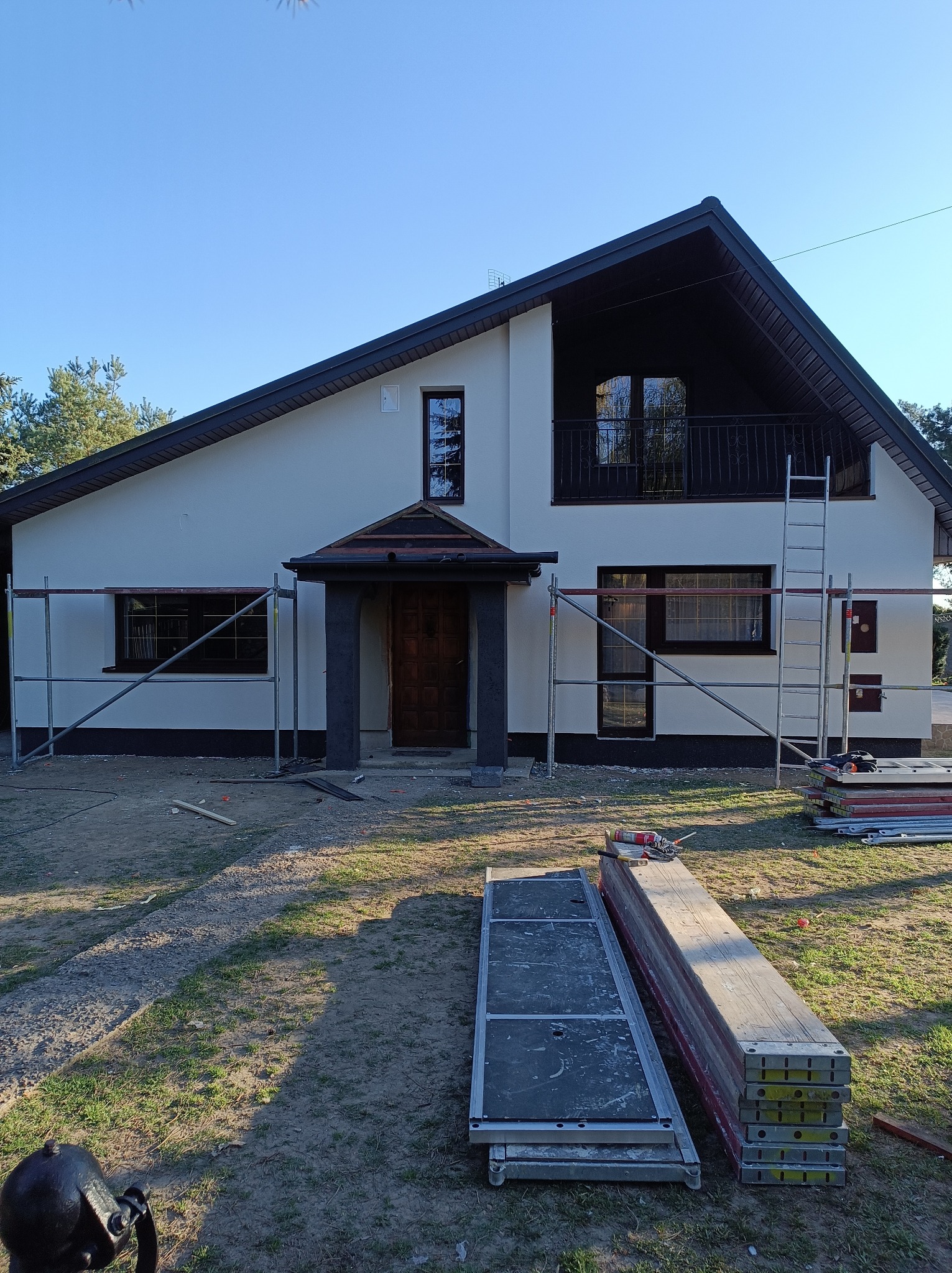 Dom jednorodzinny z białymi ścianami i ciemnym dachem w trakcie prac wykończeniowych elewacji, widoczne rusztowania, deski i narzędzia na trawniku.