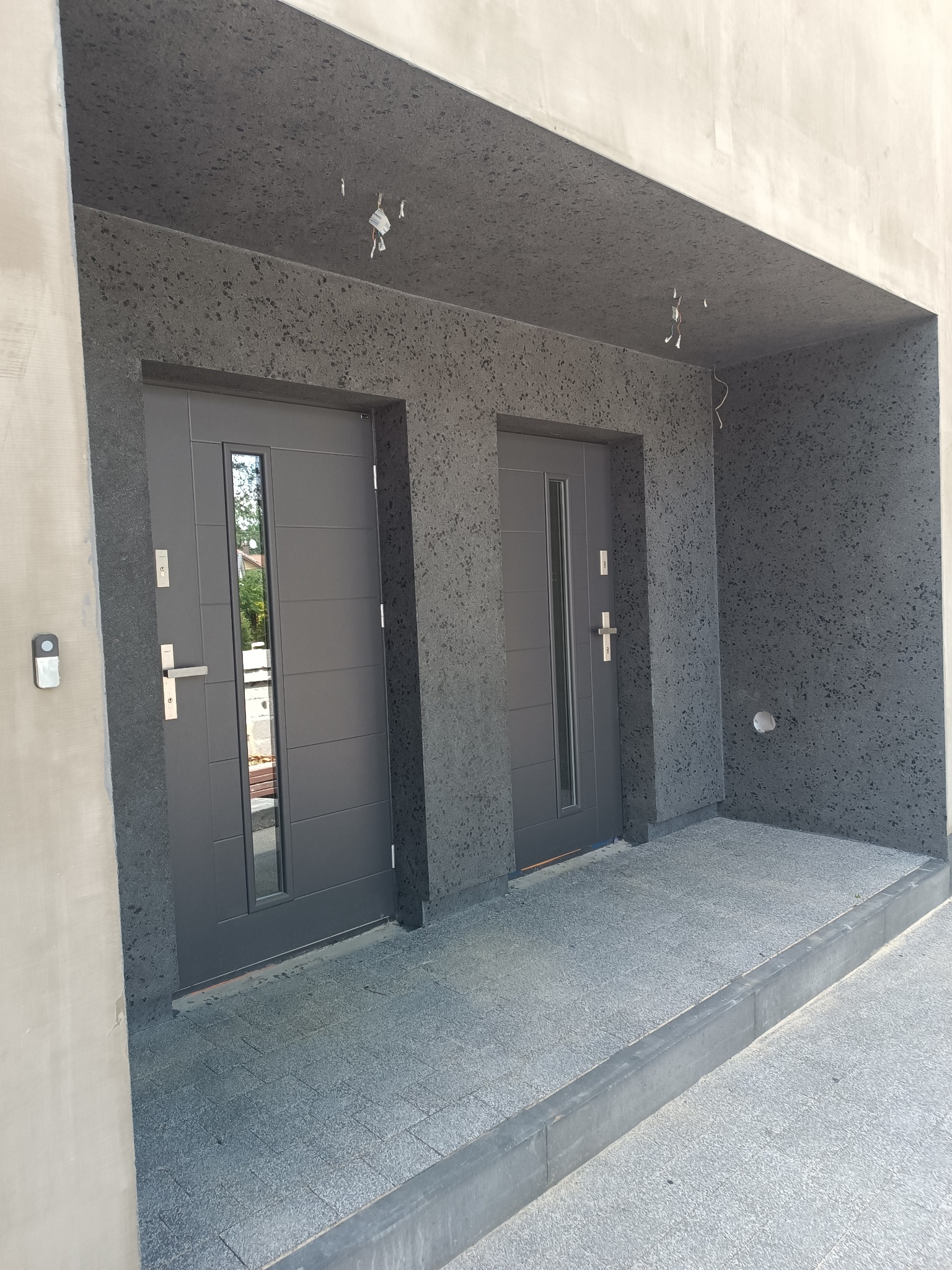 Dwa szare, nowoczesne drzwi wejściowe z przeszkleniami, osadzone w elewacji o fakturze baranka, z widocznymi przewodami elektrycznymi pod sufitem.
