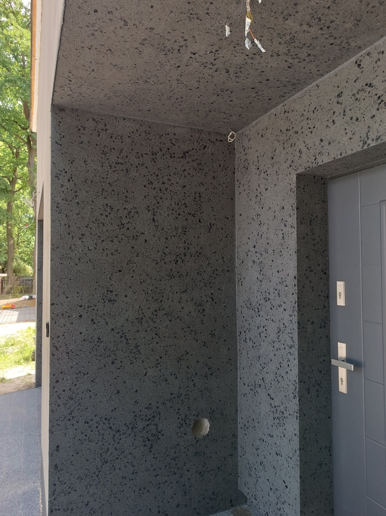 Szara elewacja zewnętrzna z efektem kamyczków, widoczne fragmenty instalacji elektrycznej i drzwi wejściowe.