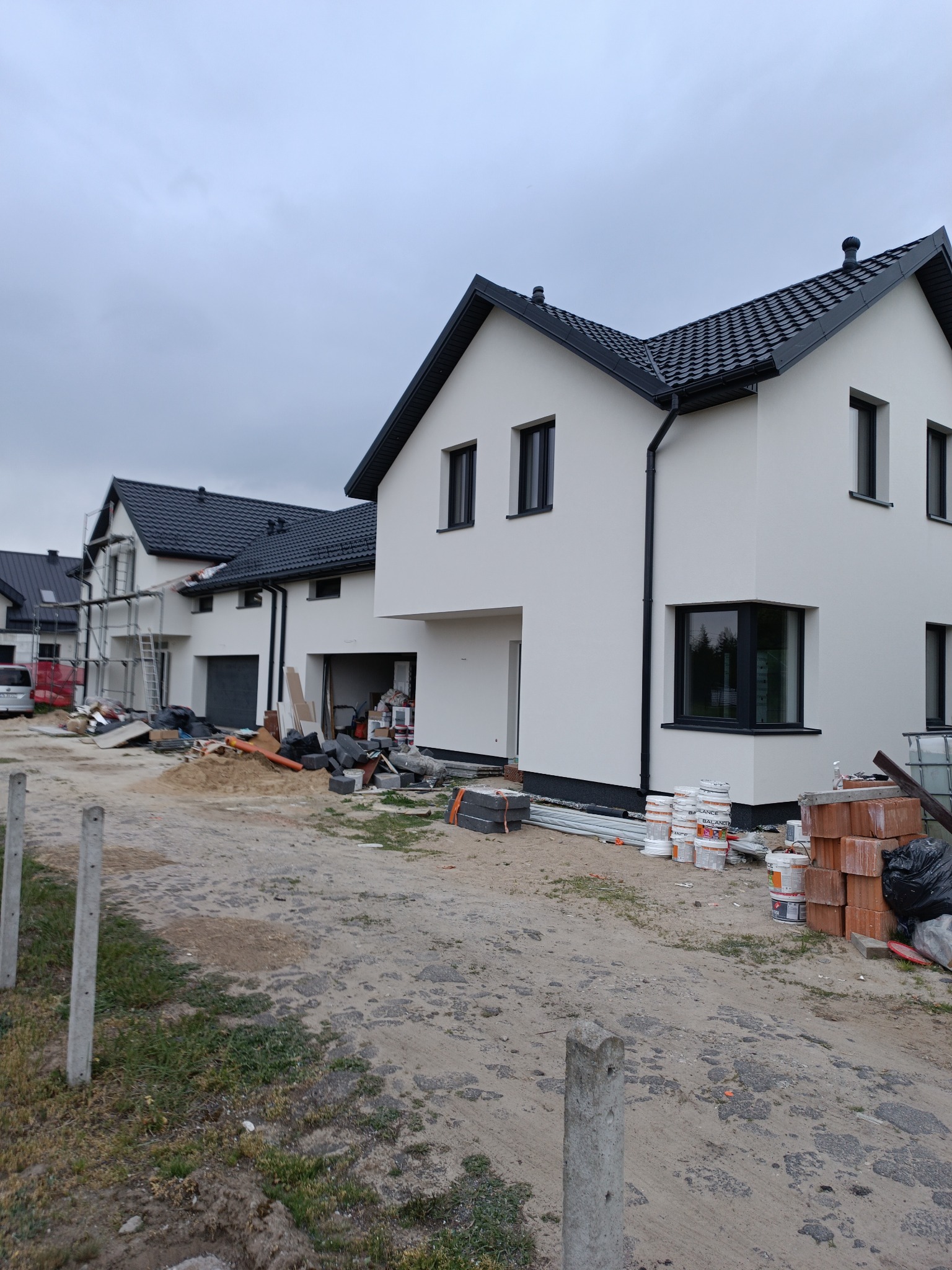 Nowoczesny dom z białą elewacją i czarnym dachem w trakcie wykończenia. Widoczne materiały budowlane i rusztowania przy sąsiednim budynku. Pochmurne niebo w tle.