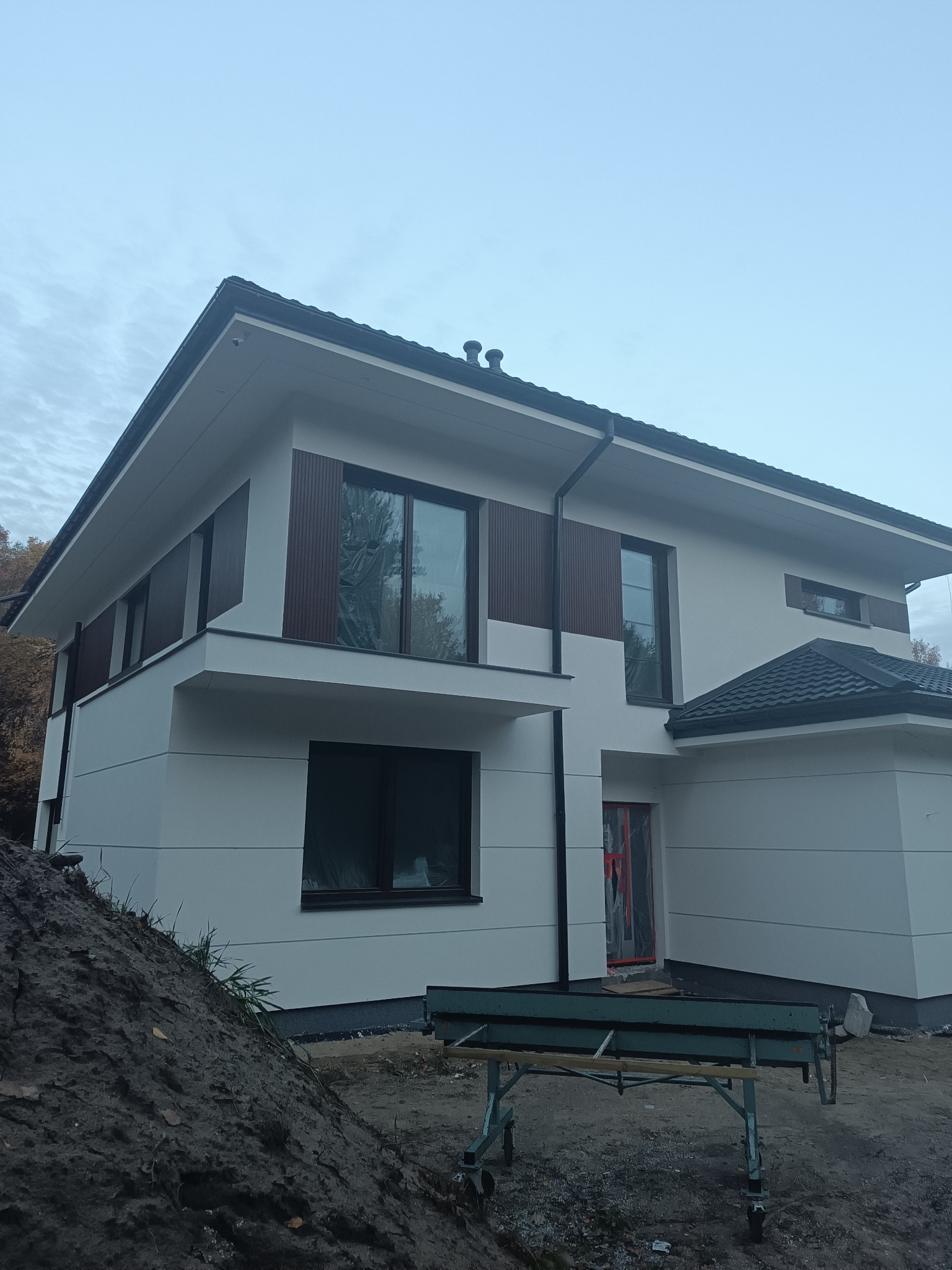 Nowoczesny dom z białą elewacją i brązowymi panelami dekoracyjnymi. Widoczne narzędzia budowlane sugerują prace wykończeniowe. Ujęcie z dołu, pochmurne niebo.