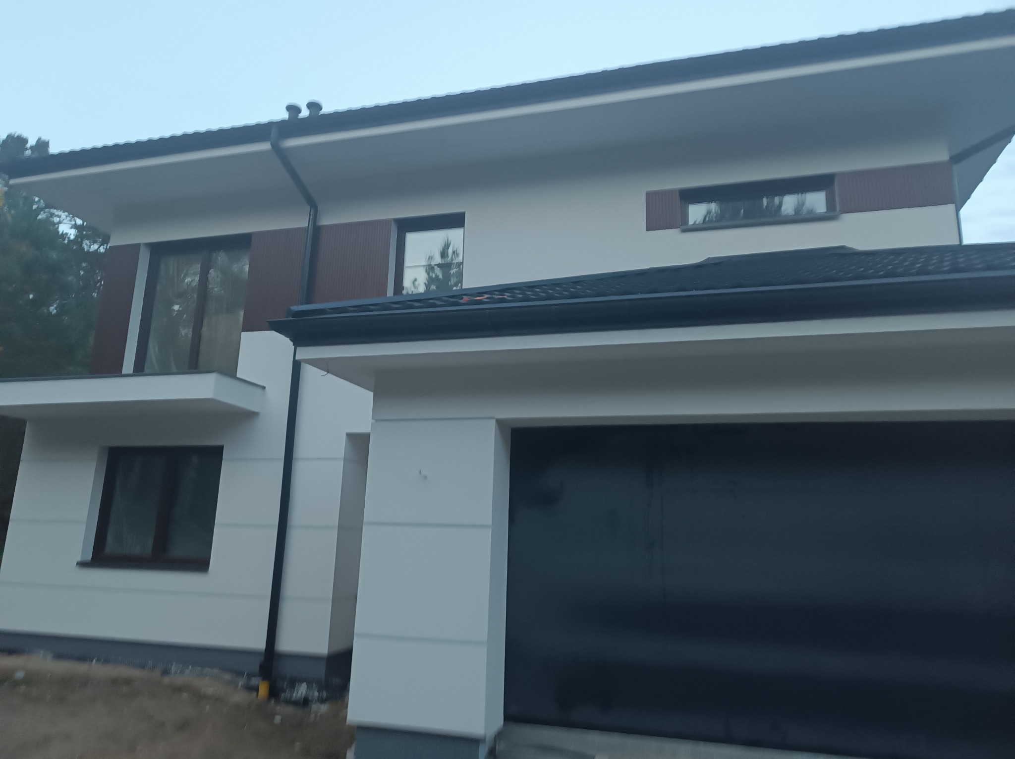 Nowoczesny dom z białą elewacją i ciemnymi akcentami, widok z dołu, garaż z bramą segmentową. Elewacja z elementami dekoracyjnymi imitującymi drewno.