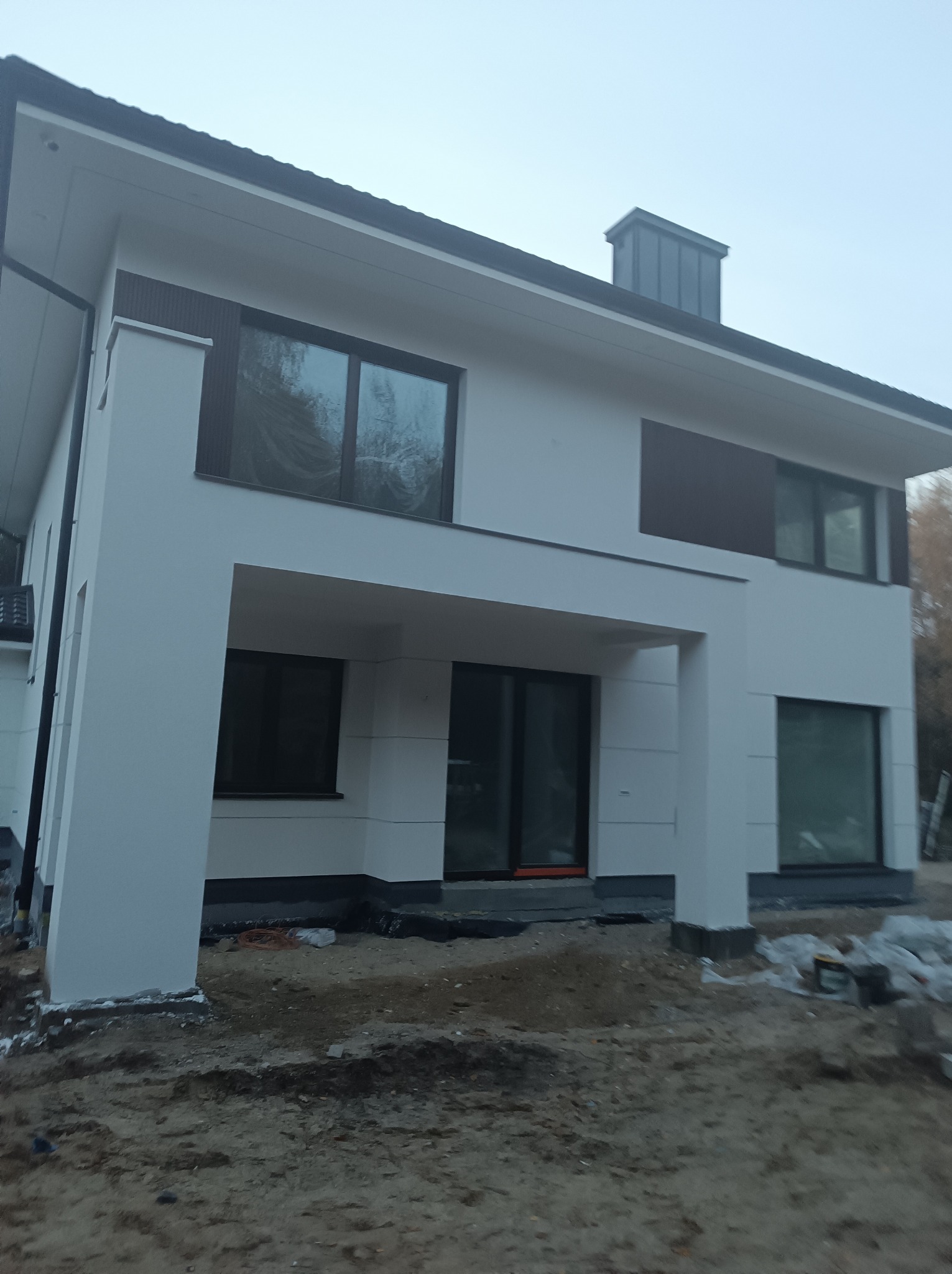 Nowoczesny dom z białą elewacją i brązowymi akcentami, widoczny w pochmurny dzień. Teren wokół domu w trakcie prac wykończeniowych, z widoczną ziemią i materiałami budowlanymi.