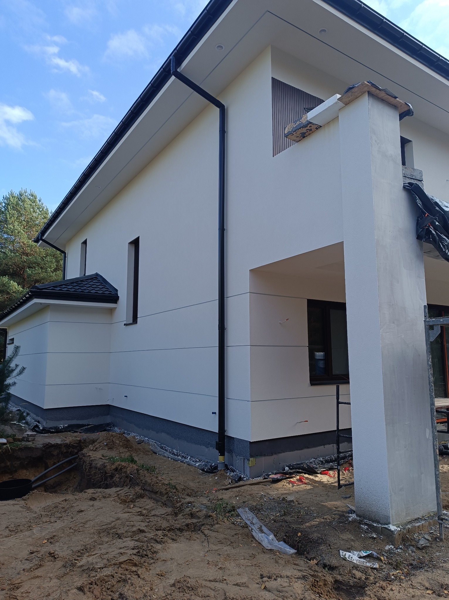 Nowoczesny dom z jasną elewacją, widoczne detale wykończenia, czarne rynny i okna. Częściowo wykończony filar. Wykop ziemny w pobliżu budynku.