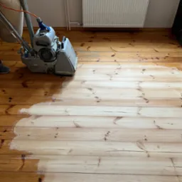 Parkiet z sosnowych desek częściowo wyszlifowany maszyną do cyklinowania, widoczny kontrast między surowym drewnem a wykończoną powierzchnią, w tle grzejnik.