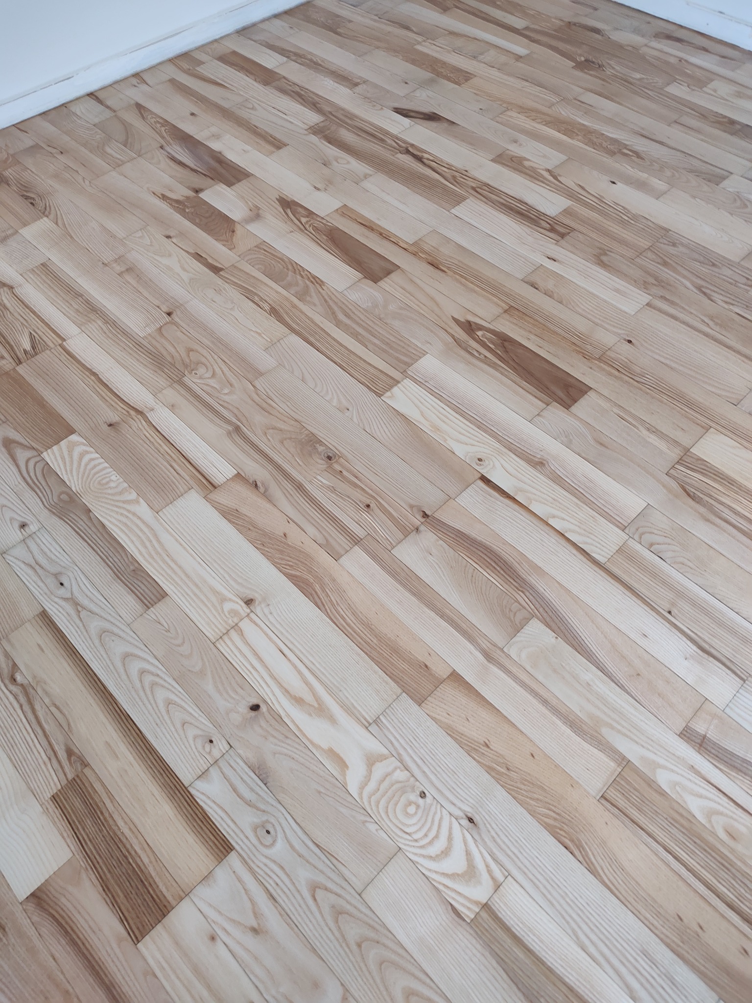 Jasne, drewniane deski parkietu ułożone pod kątem, widoczne naturalne słoje drewna i subtelne różnice w odcieniach, kontrastujące z białą ścianą w tle.