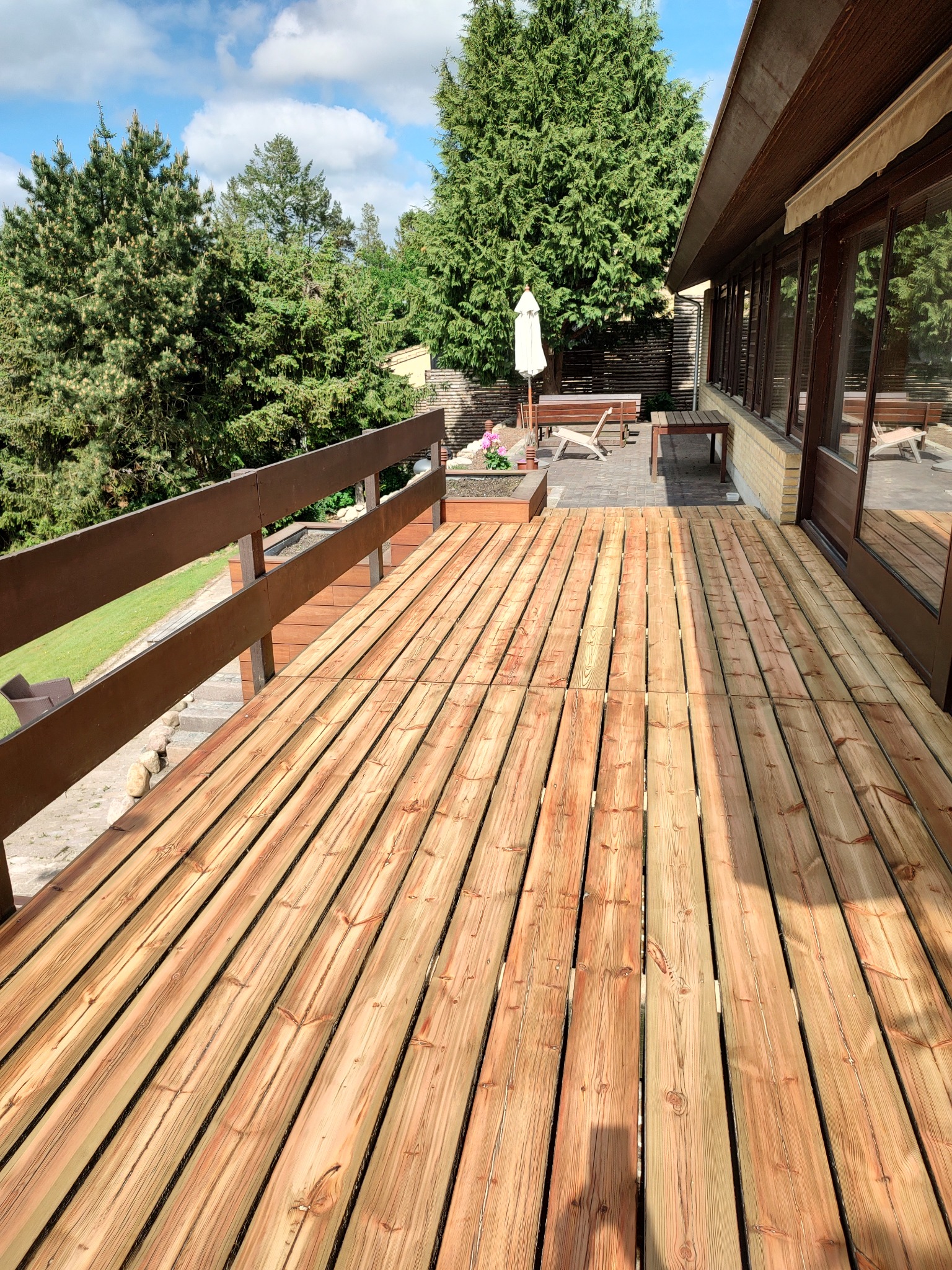 Nowo ułożony taras z desek kompozytowych w odcieniach jasnego drewna, widok z góry, z balustradą po lewej stronie i patio z meblami ogrodowymi w tle.