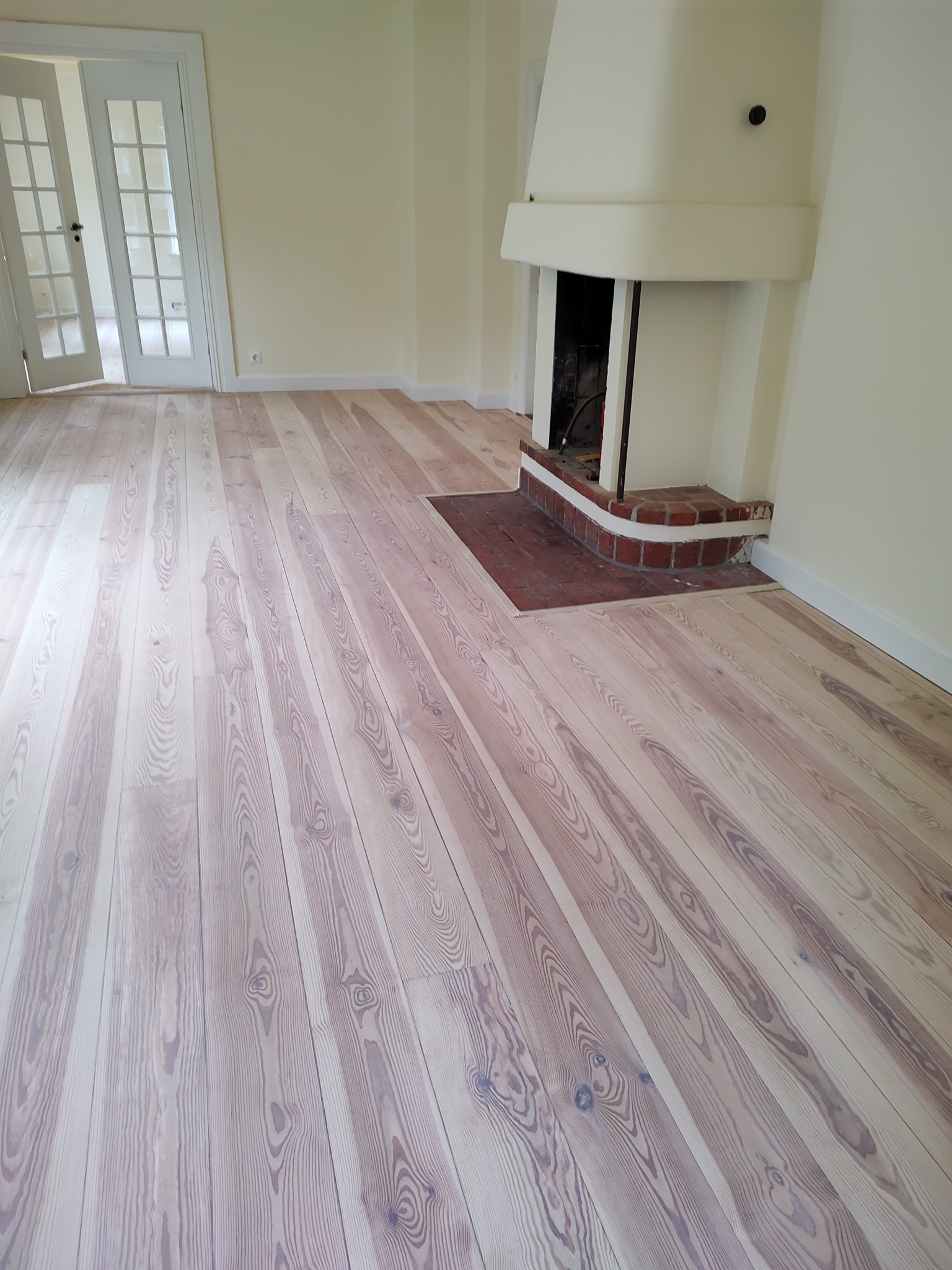 Jasne, odnowione deski podłogowe z widocznym rysunkiem drewna, ułożone w kierunku kominka z ceglaną podstawą; w tle białe ściany i drzwi.