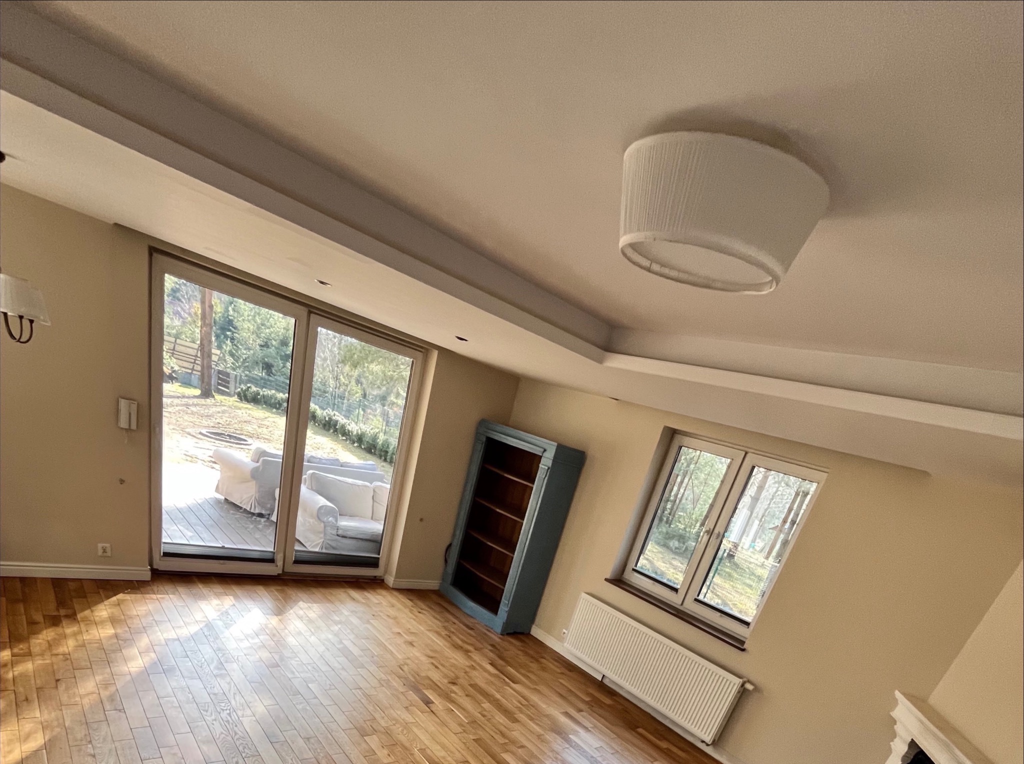 Jasny salon z parkietem ułożonym w jodełkę, widok na taras i ogród przez duże przesuwne drzwi, błękitny regał przy ścianie, biały grzejnik pod oknem.