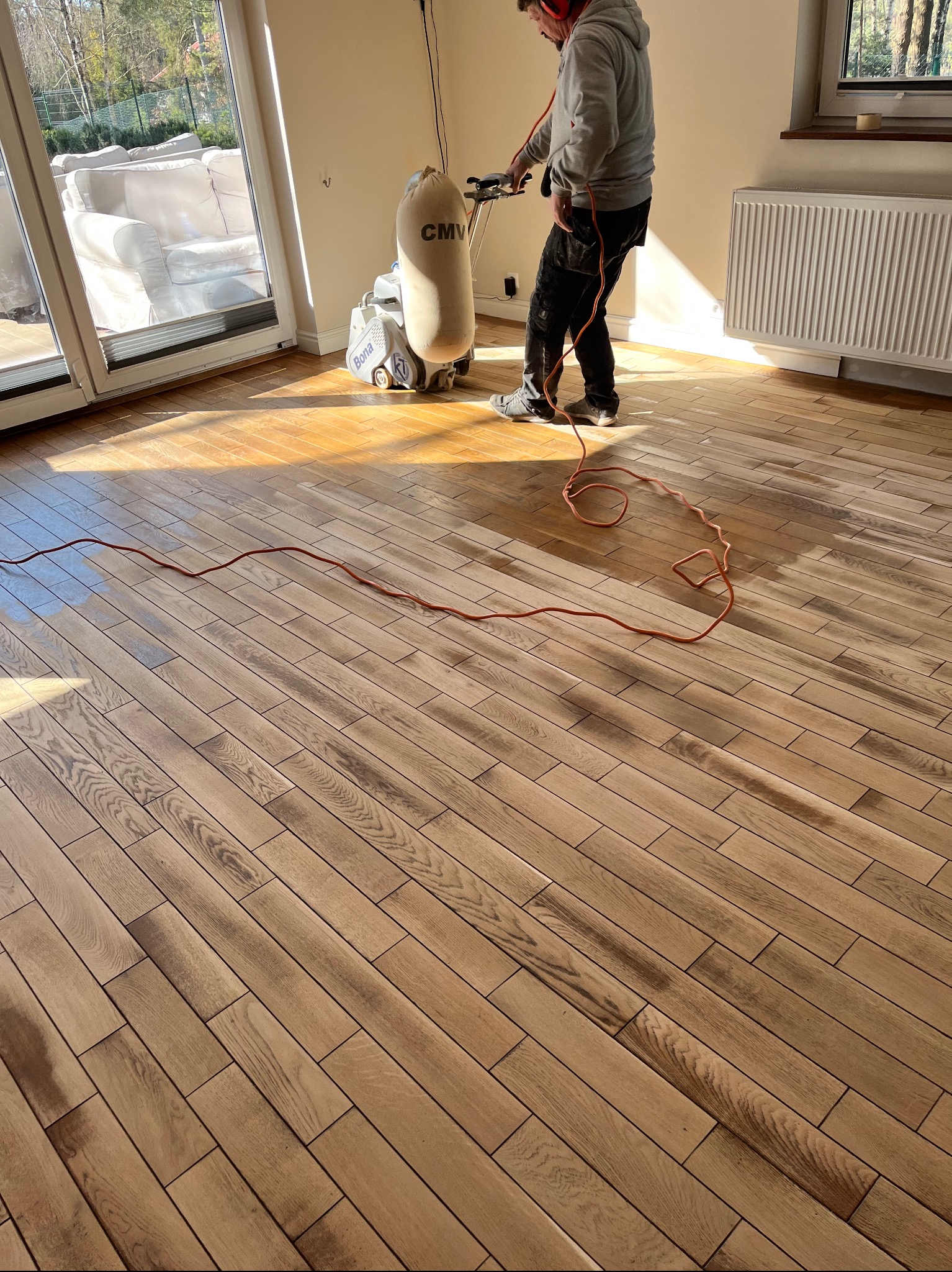 Pracownik w słuchawkach cyklinuje drewnianą podłogę parkietową maszyną Bona, widoczne ślady cyklinowania i pomarańczowy kabel zasilający